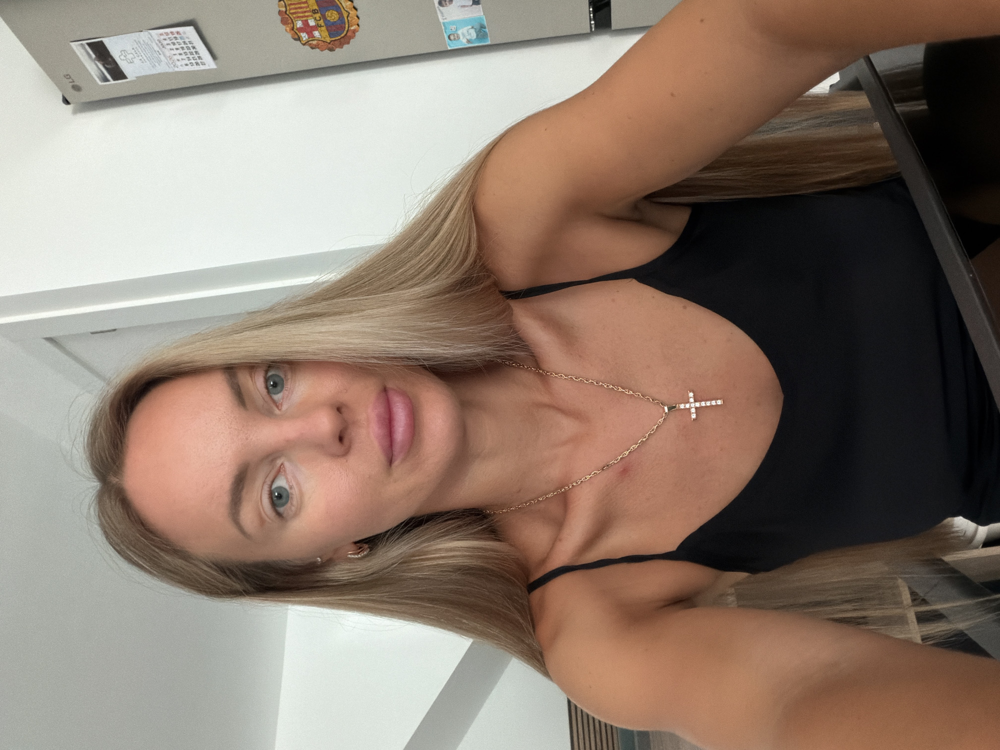 A woman with long blonde hair, wearing a black camisole and a gold cross necklace, is captured in a close-up, front-facing selfie. Her blue eyes are looking towards the camera, and her lips are slightly parted. The background includes a white wall, a door, and a glimpse of a refrigerator with a calendar and a FC Barcelona crest magnet. The scene suggests an indoor domestic setting, possibly a kitchen or hallway in a home located in Finestrat, Spain.