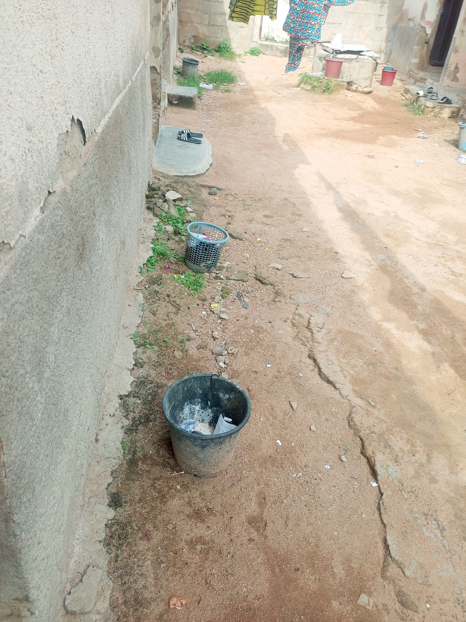 The image depicts an outdoor scene in Jalingo, Nigeria. A textured, light-colored wall runs along the left side of the frame, showing signs of wear and damage, including peeling plaster. In front of the wall, a dusty, sandy ground is visible, interspersed with patches of green vegetation and small stones.
Several objects are scattered across the ground. A black bucket filled with debris is situated near the wall in the foreground. Further back, a grey, open-weave basket, also containing some refuse, rests on the ground. To the right of the basket, a small, concrete platform is visible with a pair of black and white striped flip-flops on it. Beyond this, the ground continues, showing an uneven, worn path. In the background, parts of buildings are visible, constructed with concrete blocks. Red buckets are placed near some of these structures. Further in the distance, a person is partially visible, wearing a vibrantly patterned garment, with what appears to be laundry hanging on a line.
The lighting suggests it is daytime, with no strong shadows indicating a specific time. The overall impression is of a utilitarian outdoor space, possibly a residential or communal area, with elements of everyday life and some degree of clutter. No visible text is present in the image.