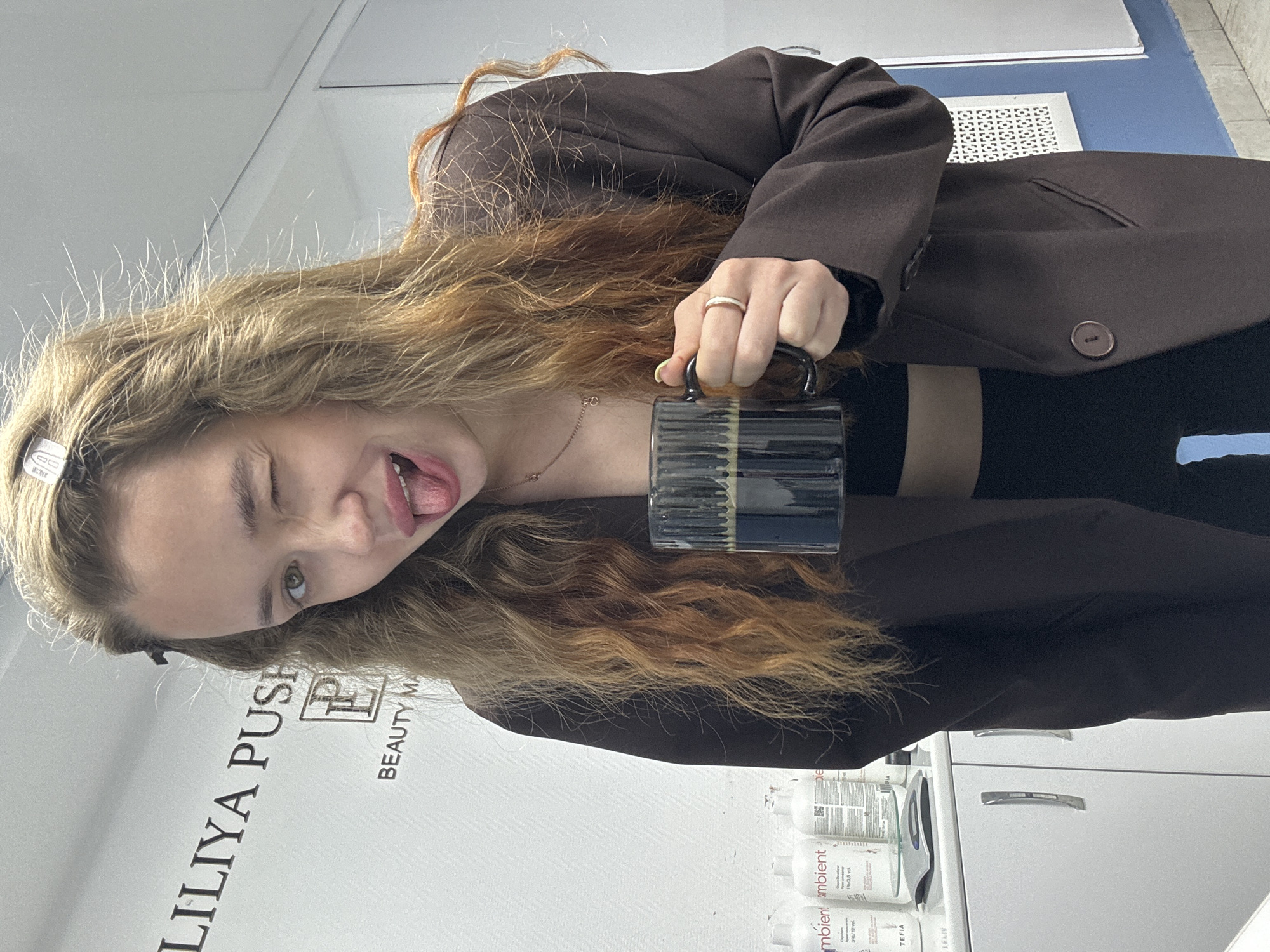 A young woman with long, wavy blonde and reddish-brown hair is captured in a playful indoor portrait in Chaikovsky, Russia. She is wearing a dark brown blazer over a black top, accessorized with a delicate gold chain necklace, a ring on her right hand, and light green/yellow nail polish. With a mischievous expression, she winks her left eye and sticks out her tongue while holding a dark, ribbed mug with a gold band.
The background suggests a beauty salon or studio. A white wall behind her features large black text that reads "LILIYA PUSH" and a "P" logo, followed by "BEAUTY MA" (likely part of "BEAUTY MASTER" or similar). To her right, a white cabinet with silver handles holds various product bottles, including some labeled "mbient ambient," "3%/3.5 vol," "3%/10 vol," and "TEFIA," alongside a scale. A blue surface and a white grate are also partially visible in the upper background. The lighting is bright, indicating an indoor setting.