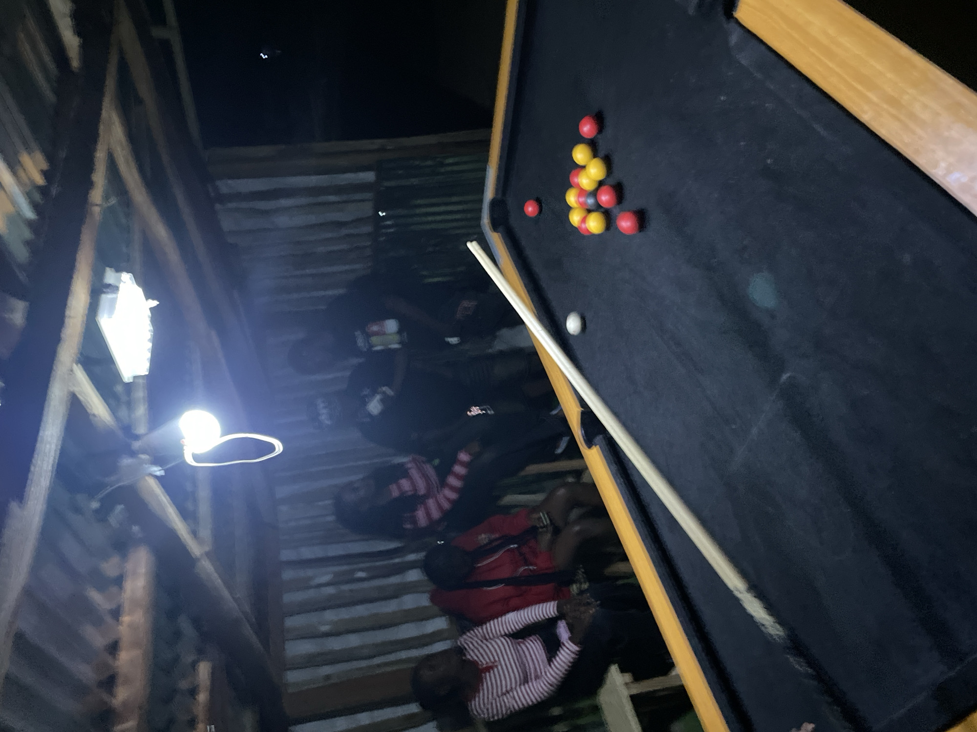 The image depicts an indoor pool hall scene in Durumi, Nigeria, under dim lighting conditions, possibly at night. The setting appears to be a rustic, open-air structure with a corrugated metal roof supported by wooden beams and a wall constructed of vertical corrugated metal sheets. A bare light bulb hangs from the ceiling, providing illumination.
In the foreground, a black pool table with a wooden frame is visible. Several pool balls, including red and yellow ones, are scattered on the table, along with a white cue ball and a cue stick resting diagonally across the table.
In the background, a group of four individuals, appearing to be young adults, are seated or standing. Two women are seated on a bench to the left, one wearing a horizontally striped pink and white shirt and the other a red hooded jacket. Behind them, two more individuals, possibly male, are standing. The lighting casts long shadows and creates a somewhat indistinct view of the people. The overall atmosphere suggests a casual gathering or game night.