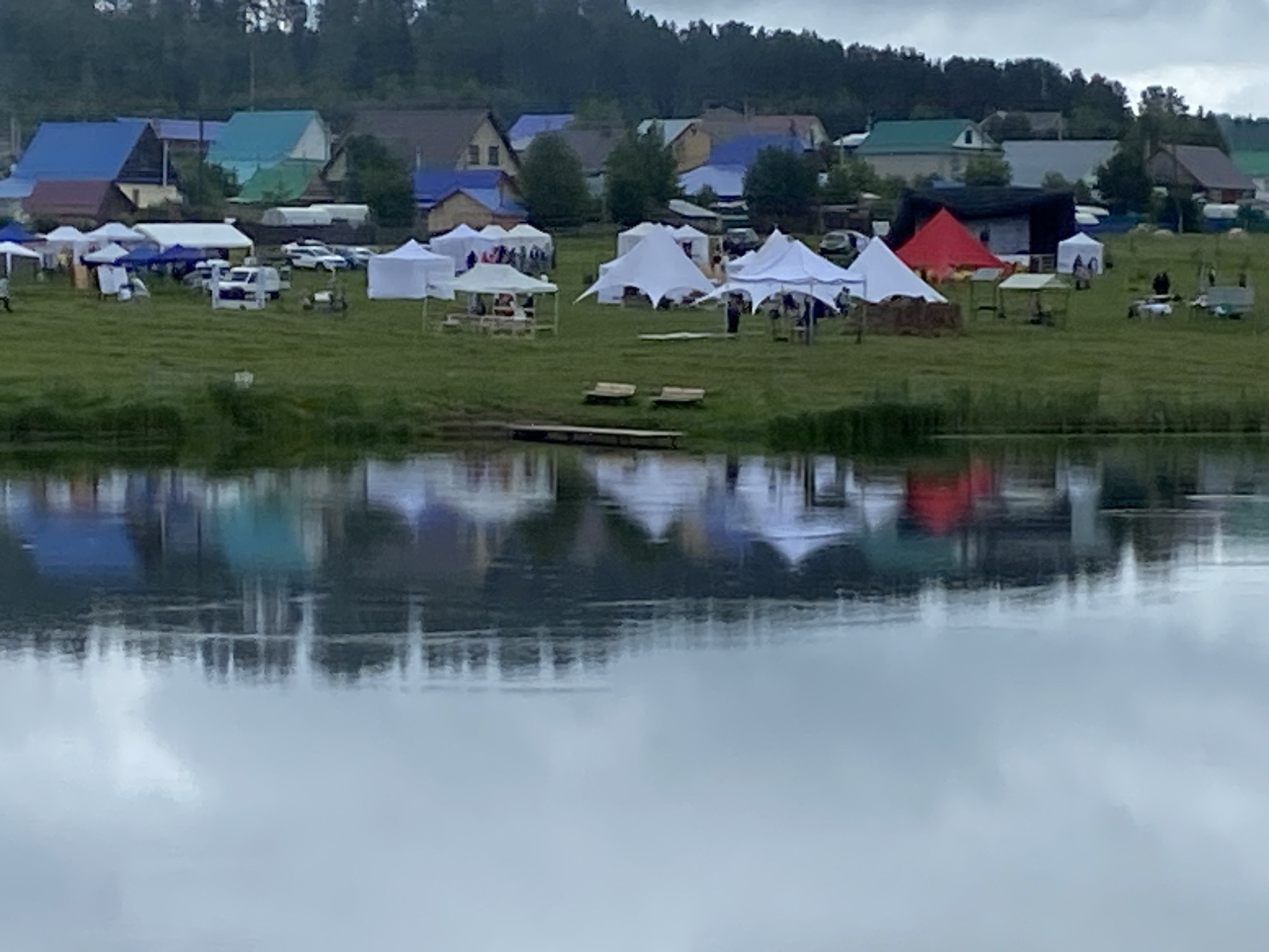 This image depicts an outdoor event taking place on a grassy field adjacent to a body of water, located in Фоки, Russia. The lower half of the frame is dominated by the calm, grey surface of the water, which reflects the overcast sky and the colors of the scene above, including blurred reflections of tents and structures.
Extending from the water's edge into the midground is a verdant grassy area. Two light-colored wooden benches are positioned near the water's bank. A small, dark-colored wooden dock or platform extends a short distance into the water.
Across the grassy field, numerous tents of varying shapes, sizes, and colors are erected, indicating an organized gathering. Prominent among these are several white canopy tents, including two large, multi-pointed, star-shaped tents. One distinct conical red tent stands out, along with other smaller tents in shades of white, blue, and green. People are dispersed among the tents, with some individuals visible near or within the tent structures, although specific activities are not discernible due to the distance. Several vehicles, including a white truck, are parked in the field between the tents. Near one tent, what appear to be hay bales or similar rustic structures are present.
In the background, beyond the field of tents, a collection of residential buildings or houses is visible. Many of these structures feature roofs in various colors, including bright blue, teal, green, and brown. A dense line of trees forms the horizon behind the buildings, suggesting a forested area. The sky above is uniformly grey and overcast.