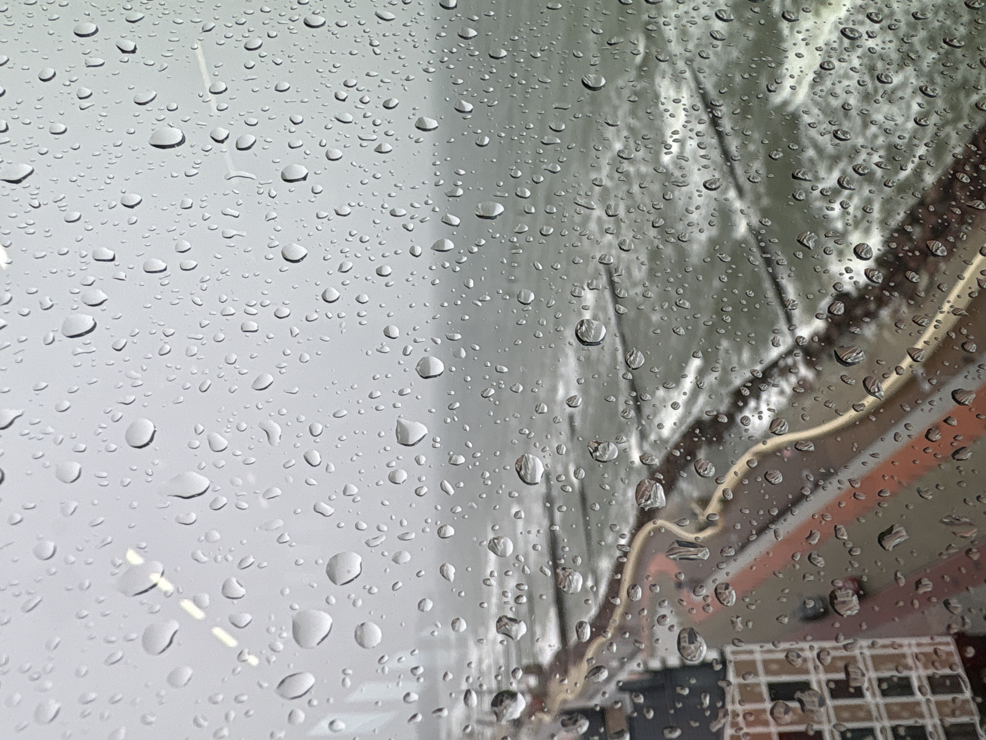 The image shows a close-up view of a surface covered in numerous water droplets. The droplets vary in size, with some being very small and numerous, while others are larger and more distinct. The surface appears to be transparent, possibly a window or glass, as a blurred background is visible through the water.
In the background, there are indistinct shapes and colors that suggest an outdoor scene. On the right side of the image, there are horizontal bands of color, appearing to be orange and brown, possibly part of a building or structure. There are also some visible electrical wires or cables in a light color. The overall impression of the background is of a somewhat overcast or muted environment, consistent with rainy weather.
The presence of the water droplets clearly indicates that it is raining or has recently rained. The light appears to be diffused, suggesting it is daytime, likely overcast. There are no people or animals visible in the image. The focus is entirely on the water droplets and the distorted view of the background through them. The scene conveys a sense of a quiet, possibly melancholic atmosphere often associated with rainy days. There is no visible text in the image.
