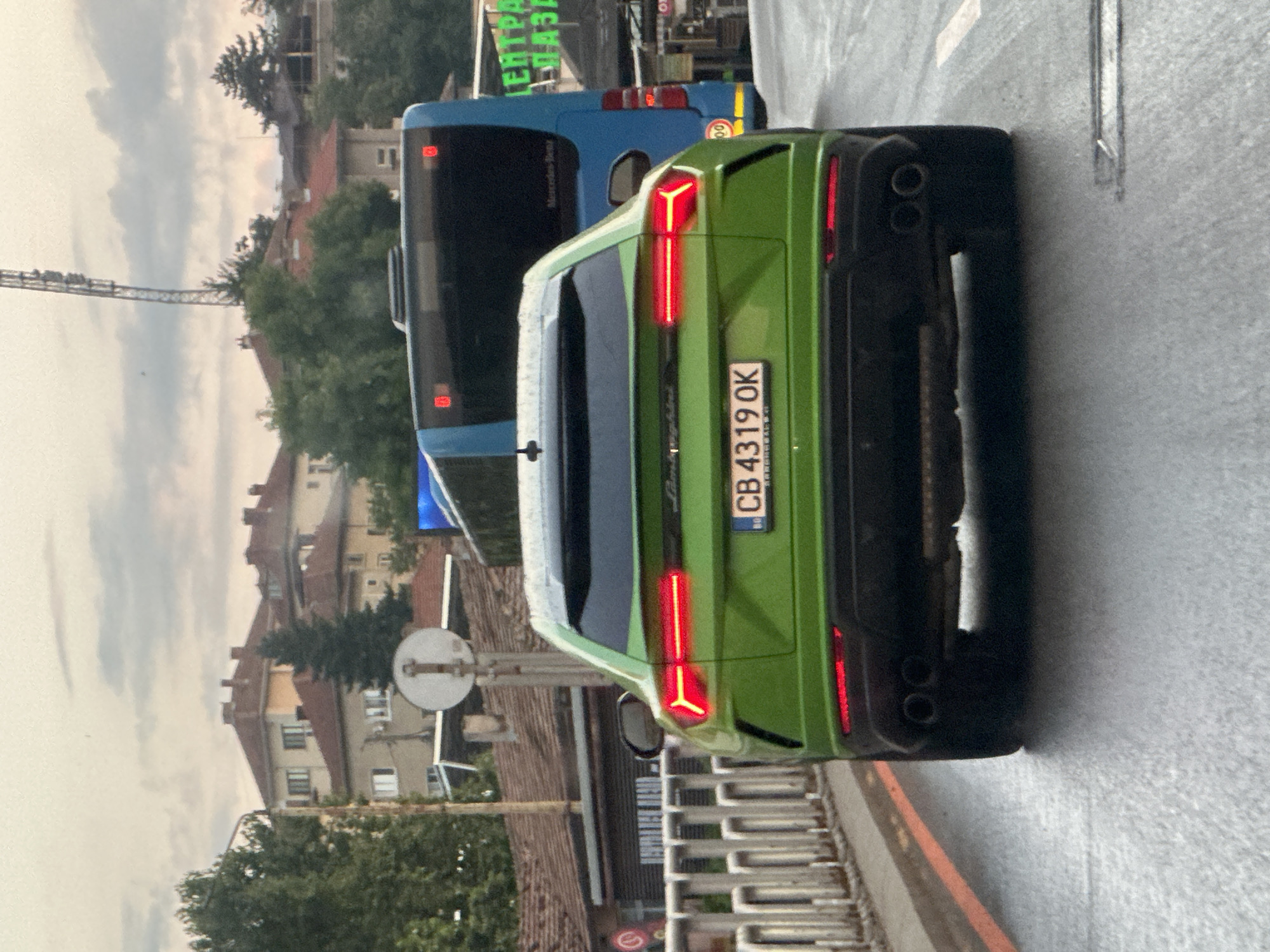 The image shows a green Lamborghini Urus parked on a wet street in Veliko Tarnovo, Bulgaria. The car's distinctive Y-shaped taillights are illuminated. A blue bus is parked behind the Lamborghini, and its rear lights are also visible. In the background, there are buildings with red tile roofs and trees. The sky is cloudy, suggesting it might be an overcast day. The street appears wet, possibly from recent rain. A metal railing and a traffic sign are also visible. The license plate of the Lamborghini reads "CB 4319 OK". The overall scene depicts a street view with vehicles and urban architecture.