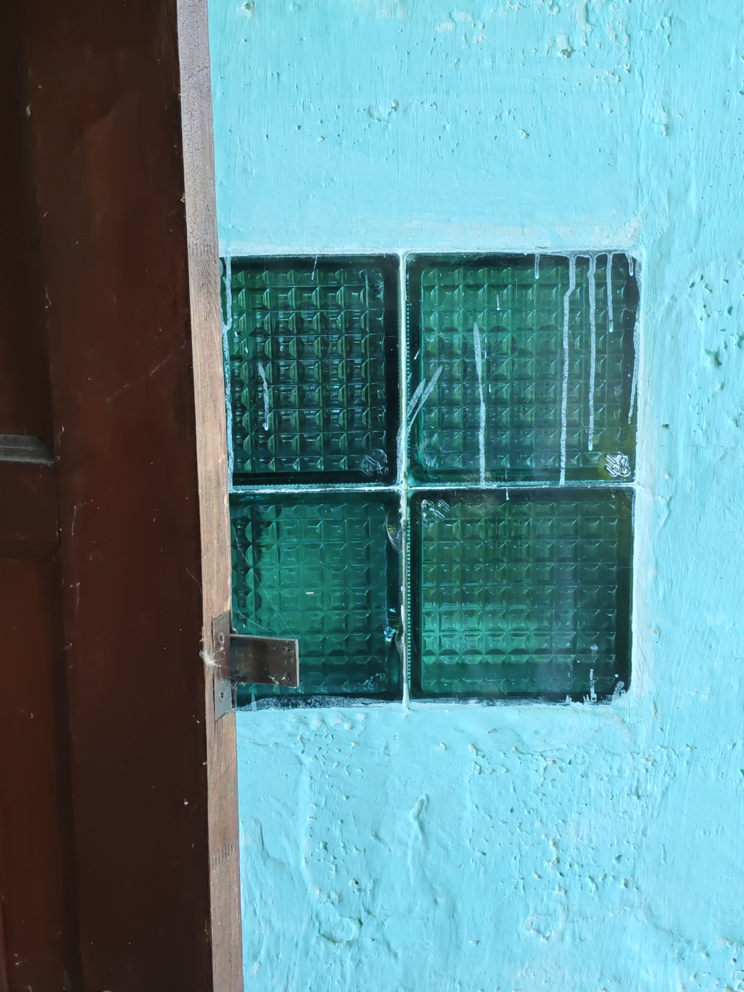 The image shows a partially open wooden door on the left, revealing a dark brown interior. To the right of the doorframe, a textured, light blue wall is visible. Embedded within this wall is a small window composed of four square, green glass blocks arranged in a 2x2 grid. The glass blocks have a patterned surface. White paint streaks, possibly from a previous painting or repair, run down the wall from the top of the window. The scene appears to be outdoors, possibly in an urban setting given the mention of Rostov-on-Don, Russia. No people are present in the image.