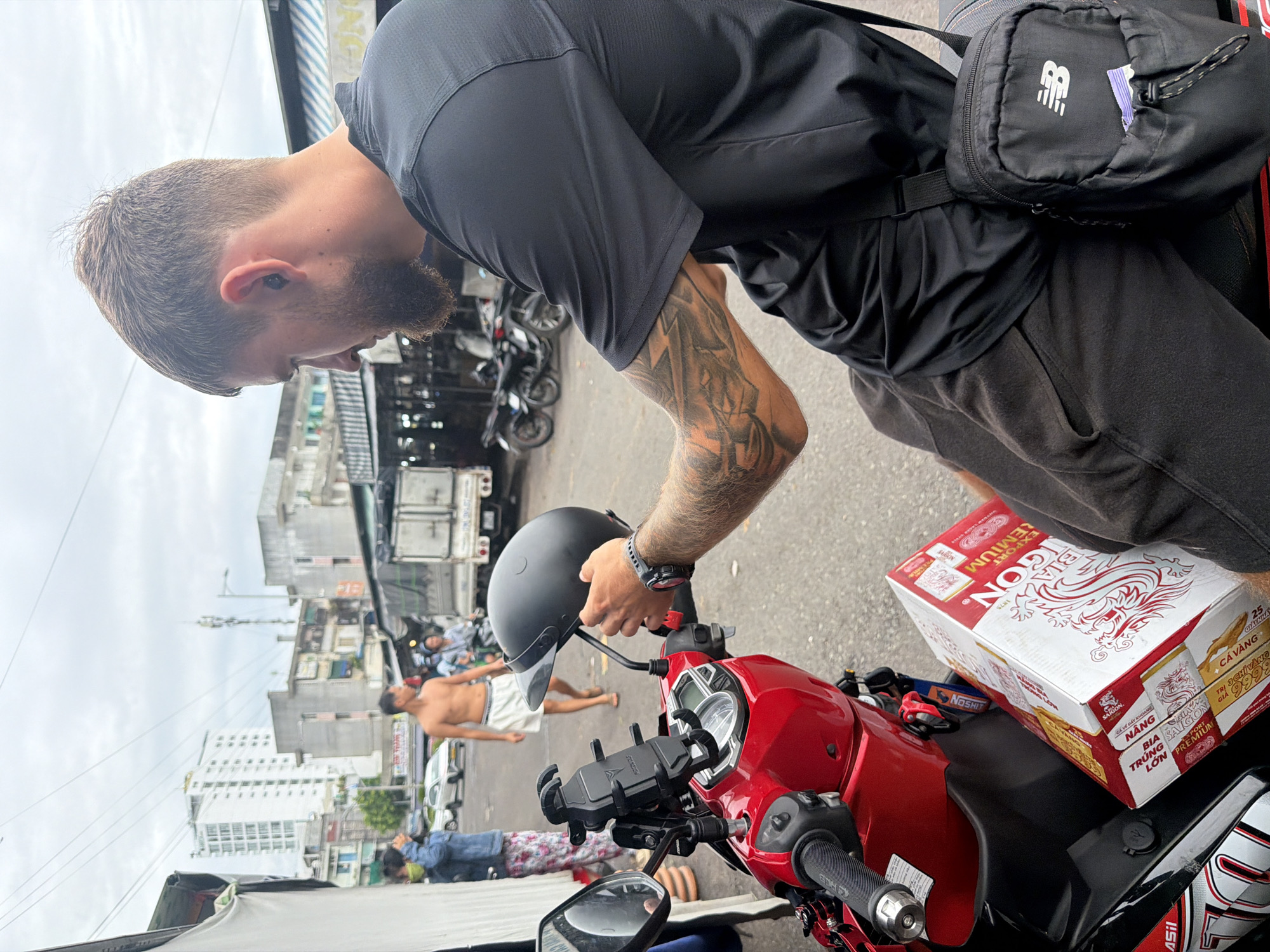 A high-angle medium shot shows a man with a beard and tattoos wearing a black shirt, placing a black helmet on a red moped. A case of "BIA SAIGON" is on the moped's seat. In the background, a shirtless man wearing white shorts walks along the street. Buildings and overhead cables indicate an urban setting, which can be identified as Nha Trang Ward, Vietnam, based on the beer brand label.