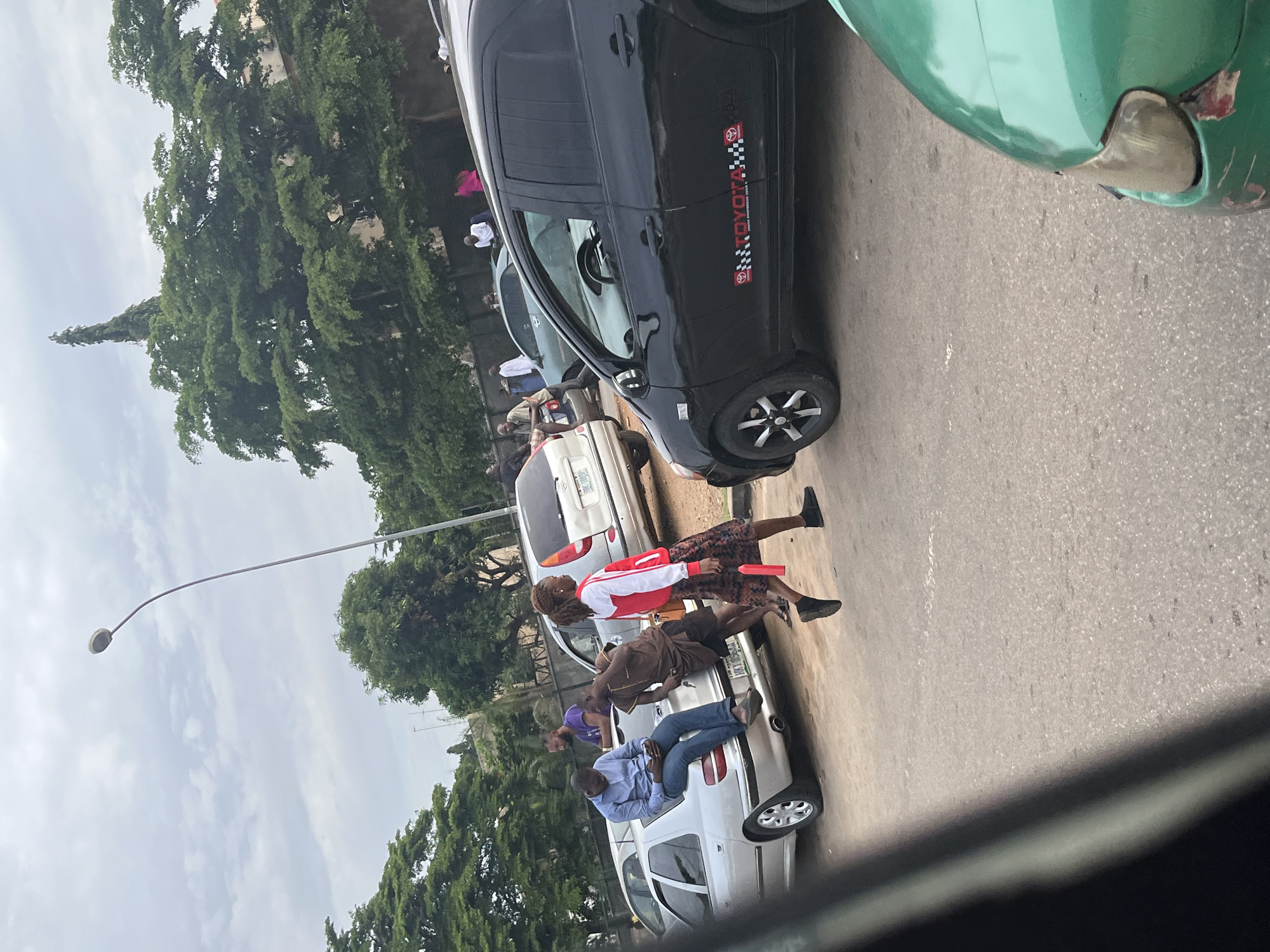 The image depicts an outdoor scene in Aso, Nigeria, likely on a street or parking area, under a cloudy sky. In the foreground, the front right quarter of a green car is visible. Behind it, a black sedan with "TOYOTA" branding and checkered accents along its side is parked, facing right. Two individuals are interacting near a silver sedan parked on the left. One, a man in a blue collared shirt and jeans, is seated on the trunk of the silver sedan, looking at his phone. Another man, wearing a brown traditional outfit, is standing next to him, leaning on the car. A woman in a red and white jacket and a patterned skirt is walking in front of the silver sedan, moving towards the right of the frame. In the background, several trees, including a tall conifer, line the area behind the cars. Other vehicles and a few people are also present in the distance. A street lamp with a curved pole stands prominently on the left side of the image.