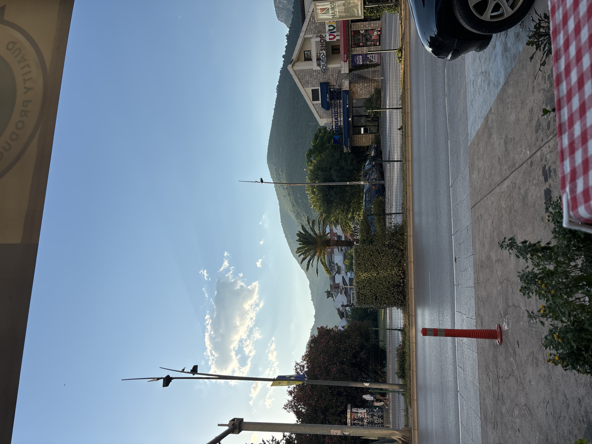 This image depicts a street scene in Budva, Montenegro, bathed in the warm light of a late afternoon sun. The sky is a clear, bright blue, with a few wispy clouds catching the sunlight. In the foreground, a red traffic cone stands on a concrete sidewalk, suggesting a temporary road closure or construction. Beyond the cone, a wet asphalt road stretches out, reflecting the sky.
On the opposite side of the road, a neatly trimmed hedge separates the sidewalk from a commercial area. Buildings line the street, their architecture a mix of modern and traditional styles. One building, constructed of stone, houses an "Adriatic Bank" and an "Auto Shop." Another building features a red awning with the word "VICTORY" visible, alongside a sign for "Namos." A palm tree stands tall near these establishments, adding a tropical touch to the urban landscape.
In the background, a lush green mountain rises, its slopes covered in dense vegetation. The sunlight casts shadows and highlights on the mountain, emphasizing its contours. A few cars are parked along the street, and a couple of people can be seen walking on the sidewalk in the distance. Overhead, a light pole with multiple fixtures stands tall, and further down the street, another pole with a yellow and blue flag is visible. A checkered tablecloth is partially visible in the lower right corner, hinting at a cafe or restaurant setting.