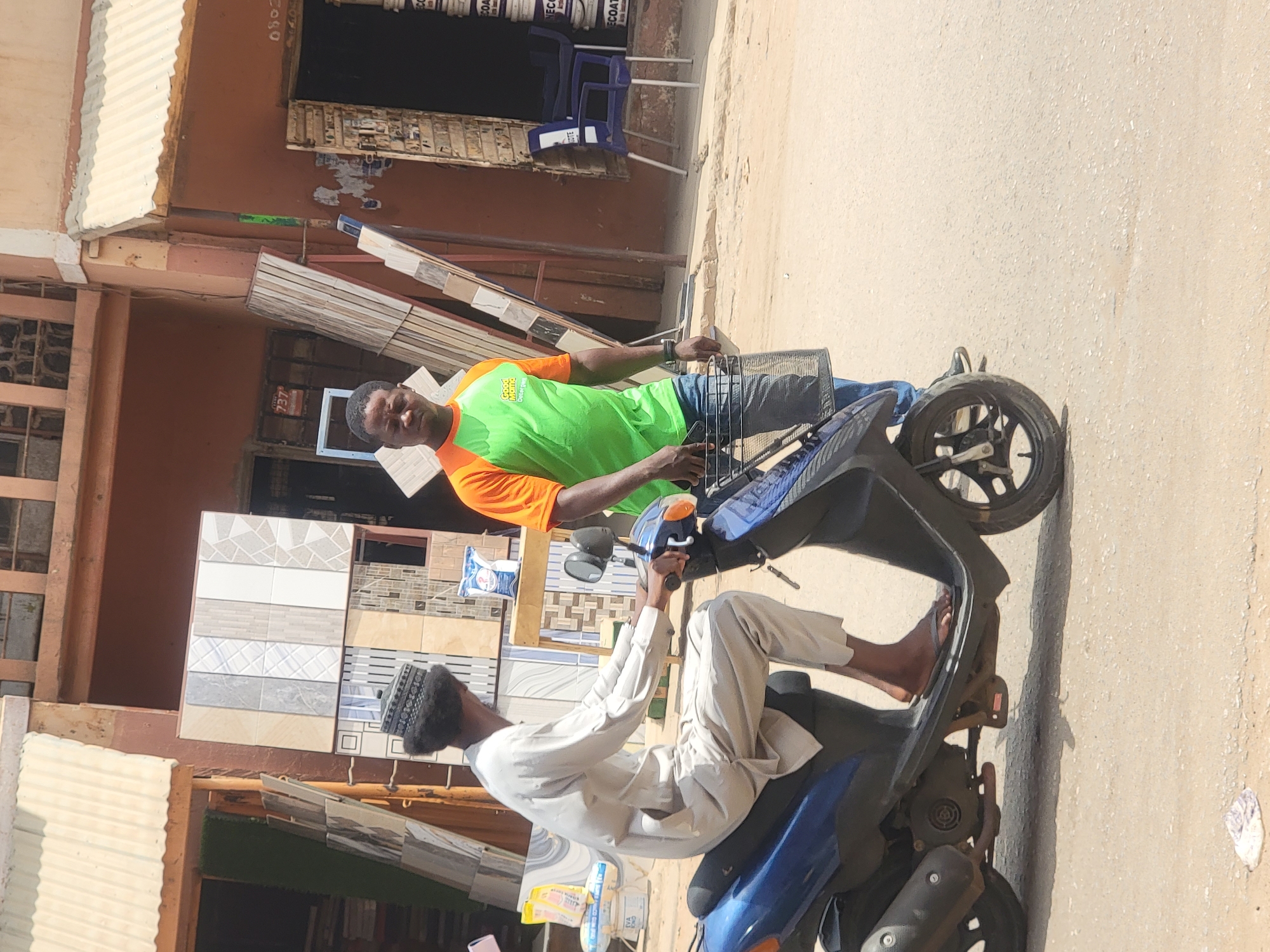 Two men stand alongside a scooter in the city of Maiburiji, Nigeria. One man, wearing a green and orange shirt and jeans, is holding onto the front of the scooter. The other man, wearing a white robe and head covering, is sitting on the scooter with his bare feet on the scooter's floorboard. Tiles are displayed in front of the men, and a building is behind them.