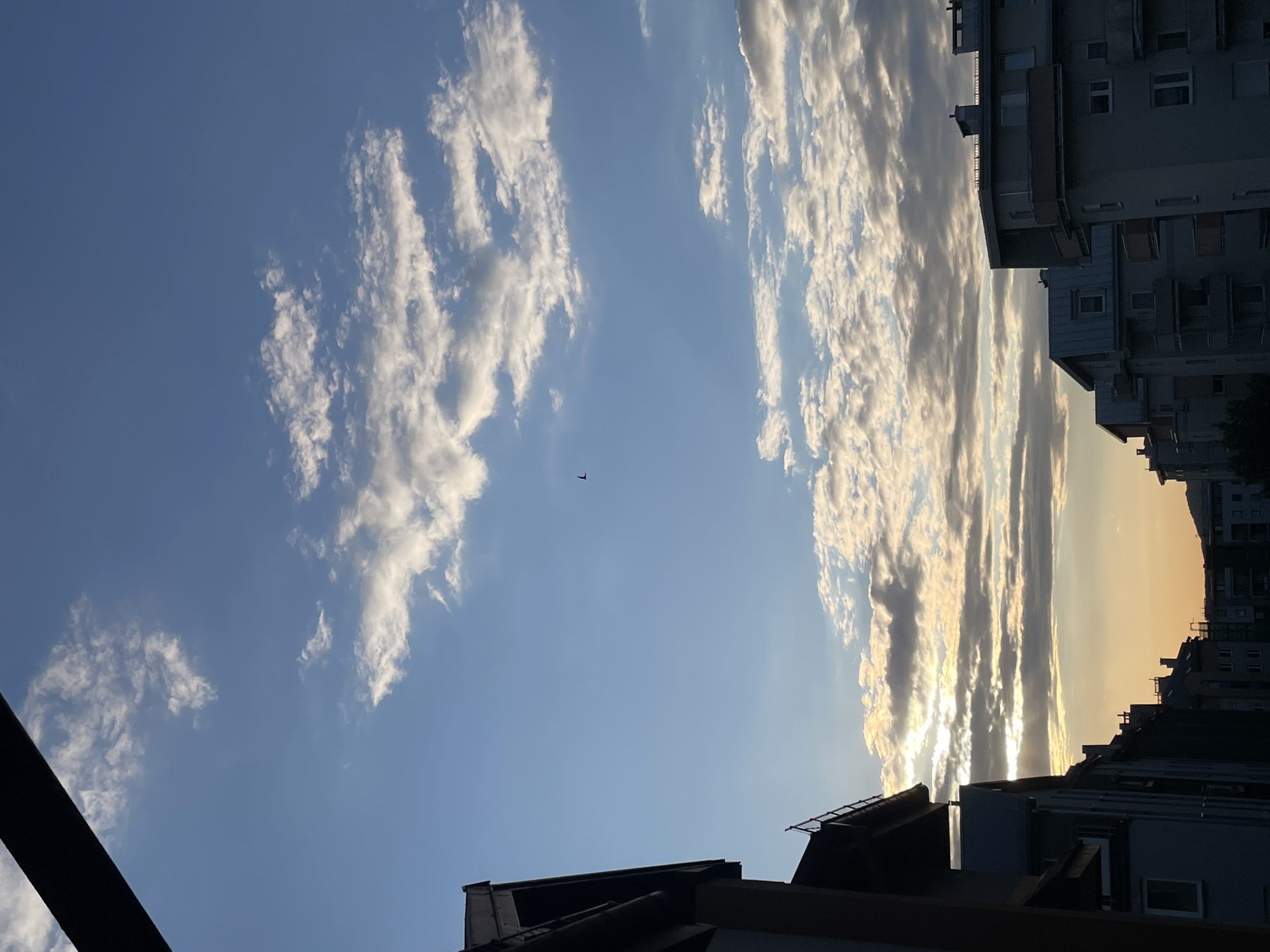 The image captures a view looking upwards towards the sky, framed by buildings in an urban environment. On the left side of the image, there is a scattering of puffy white clouds against a light blue sky. A small object, possibly a plane or bird, is visible in the center of the blue sky. To the right, the clouds are denser and more elongated, reflecting sunlight in a way that suggests the setting sun. Silhouettes of buildings with flat roofs and windows can be seen below, providing context to the urban location. The scene is a mix of natural beauty and urban landscape.