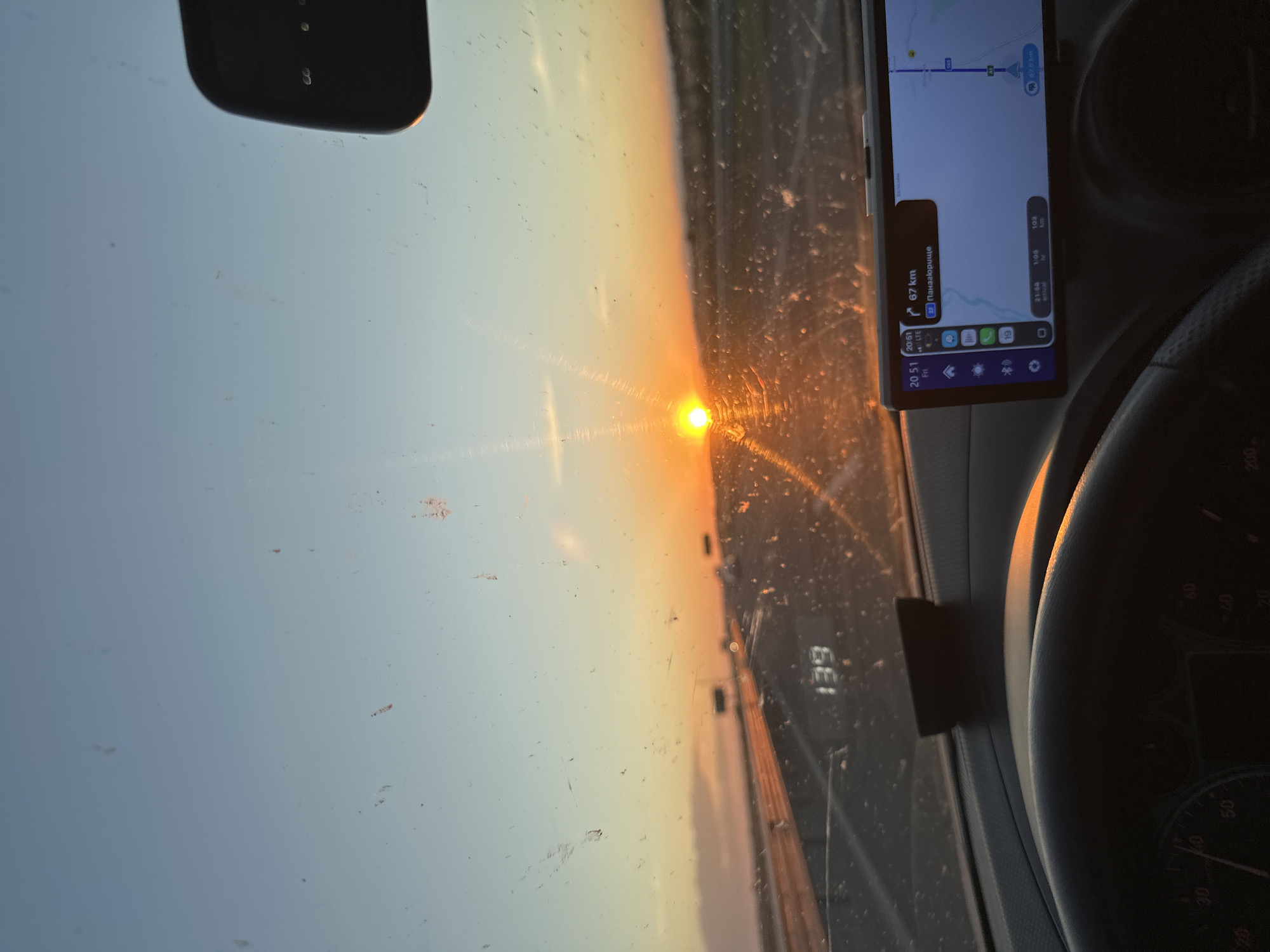 The image depicts the interior of a car looking out the windshield at sunset. The sun is low on the horizon, casting a warm orange glow across the sky and the road ahead. The road appears to be a highway with multiple lanes, and there are signs and other vehicles visible in the distance. The windshield is speckled with dirt and smudges, which are illuminated by the setting sun.
In the foreground, the dashboard and steering wheel of the car are visible. A GPS device is mounted on the dashboard, showing a map and navigation information. The time displayed on the GPS is 20:51, and the remaining distance to the destination is 67 km. The destination is labeled as "Пanaгloрице". A digital speedometer on the dashboard shows a speed of 139. The overall scene suggests a road trip during the evening, with the driver possibly heading towards the indicated destination. The location is not specified beyond "Unknown, Unknown."