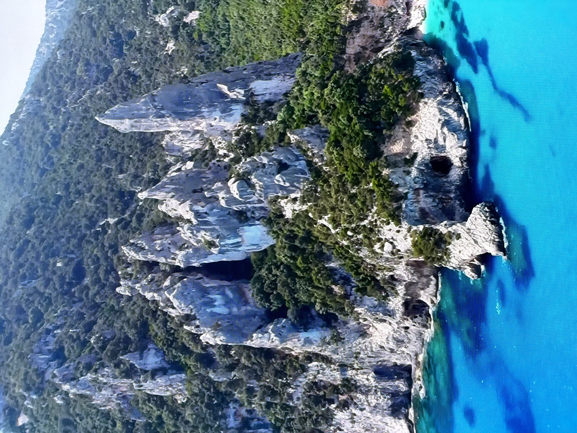 This is a high-angle aerial shot of a rugged, mountainous coastline with vibrant turquoise water. The mountains are steep and rocky, with visible caves and crevices, and are densely covered in green vegetation. The water is clear and shallow near the shore, revealing dark patches that could be rocks or seaweed. The scene appears to be a natural landscape, likely a coastal area known for its dramatic cliffs and clear waters. There are no people or man-made structures visible in the image. There is no discernible connection to the city of Chirchik, Uzbekistan, as the landscape is characteristic of Mediterranean coastlines or similar geological formations, not the geography of Central Asia.