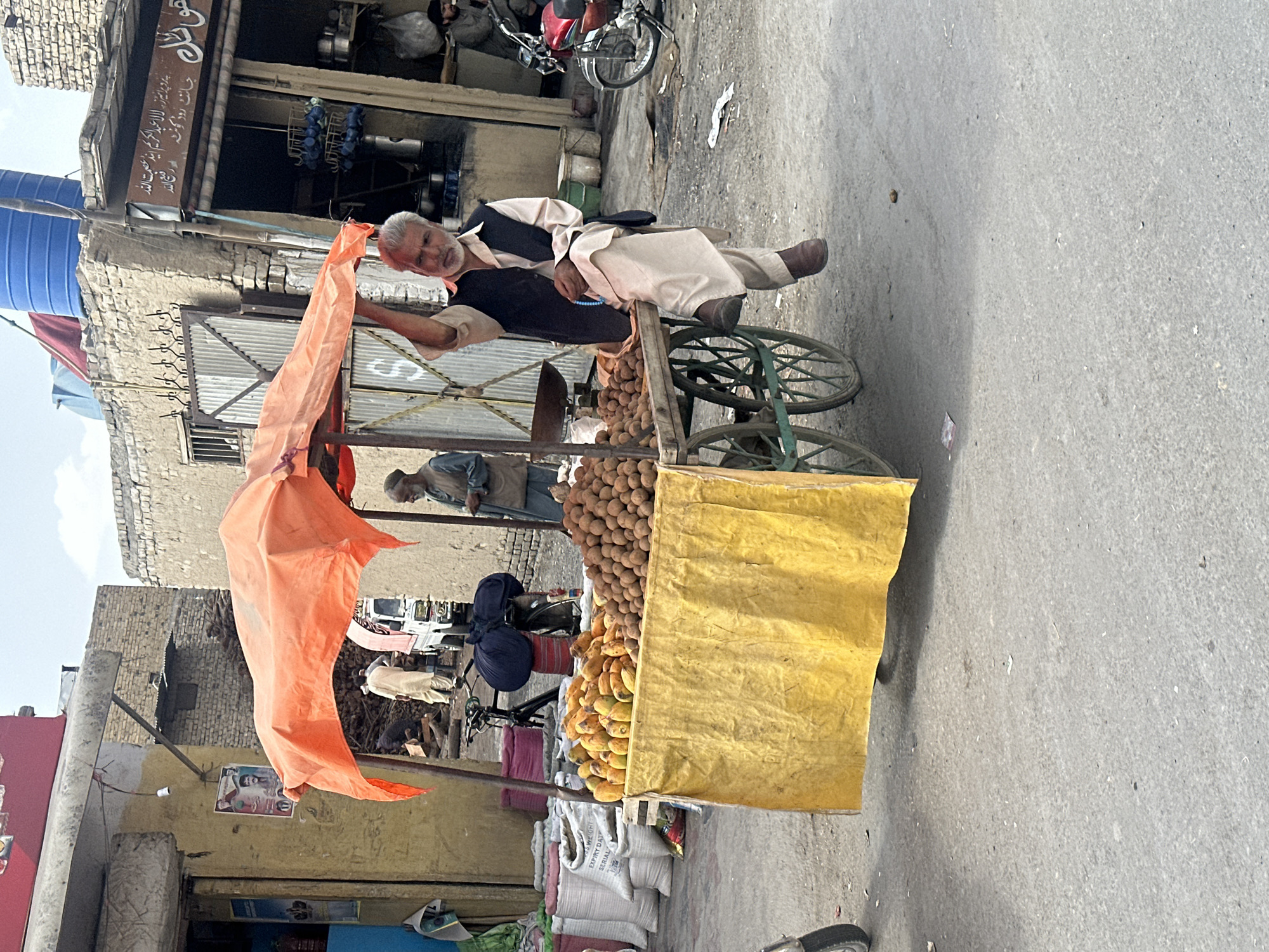 An elderly man lies on a fruit cart on a street in Quetta City Tehsil, Pakistan. The cart is stocked with brown fruits and bananas and covered with a yellow tarp, and an orange tarp is attached to the cart's corner. Behind the cart, another person stands, and two people on bicycles are further back. The scene is set against a backdrop of older buildings with signs in Urdu, and a red motorbike and various supplies are visible in the surroundings. The signage in the shop located close to the man reads: "پر پرانیز، لال عبد الکریم این عصمت الله جانٹ روڈ کوئٹہ اور رفیع الله"