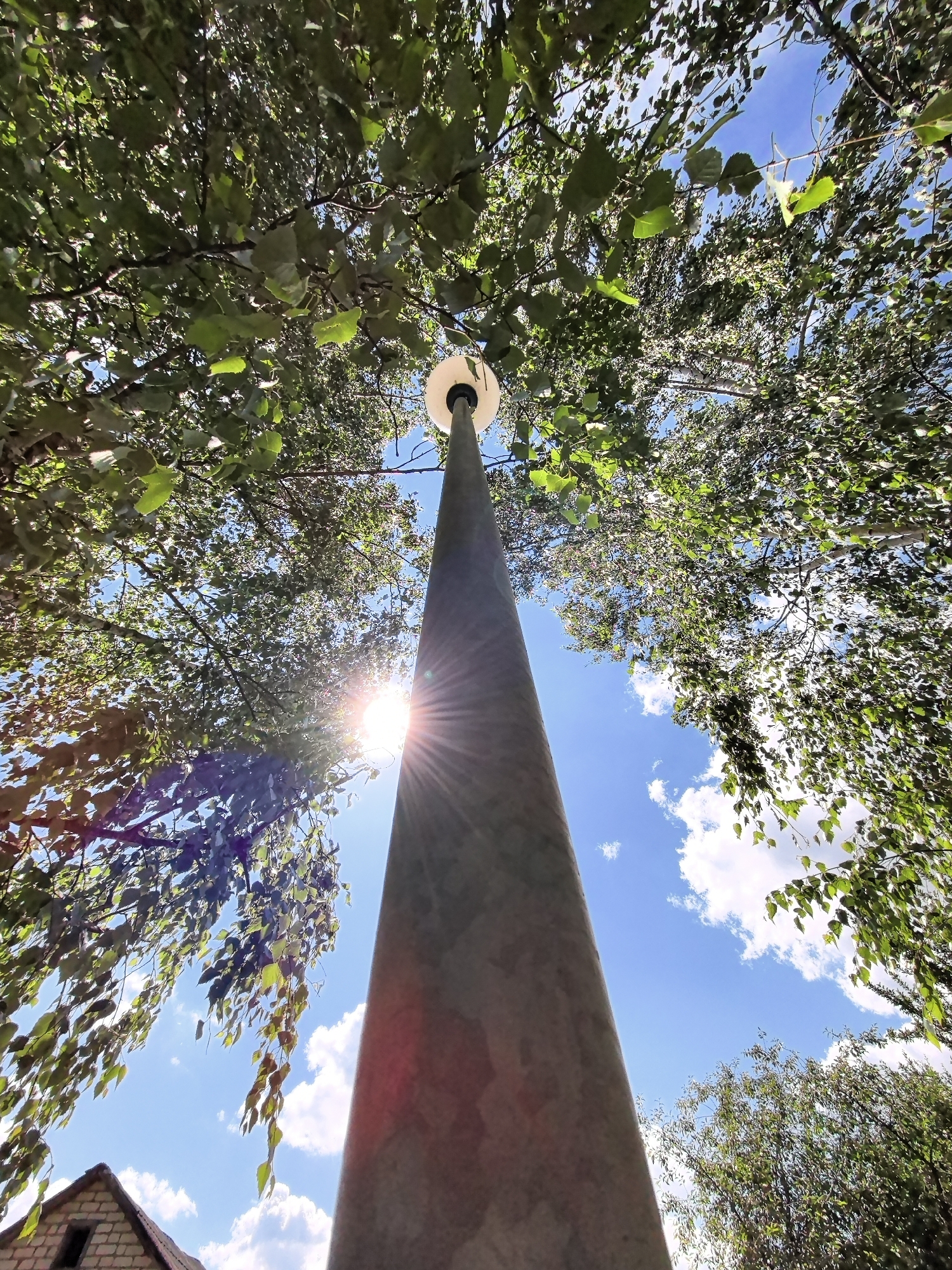 A low-angle, wide-shot photograph captures a tall, concrete lamppost extending upwards into a bright, blue sky dotted with white, fluffy clouds. The lamppost is positioned slightly off-center to the right, its conical form tapering towards a circular, white lamp fixture at its apex. Sunlight flares from behind the lamppost, creating a lens flare effect that radiates outwards. Lush green foliage from overhead trees drapes down around the lamppost, framing the sky. The leaves are vibrantly green, suggesting a warm, sunny day. In the lower left corner of the frame, a small portion of a brick building with a slanted roof is visible. The scene appears to be outdoors, possibly in a park or residential area, with a clear emphasis on the interplay between the artificial structure, natural elements, and the sky.