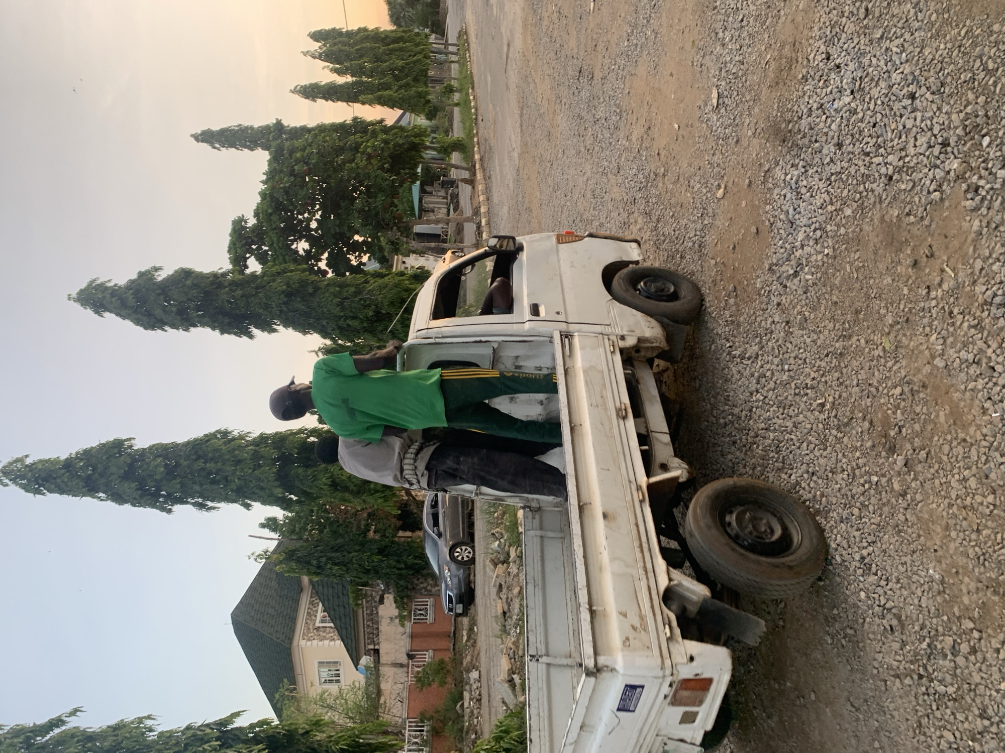 Two men stand on the bed of a white flatbed truck. One man wears a green shirt and black pants, and the other wears a gray shirt and black pants. The truck is parked on a gravel surface. In the background are trees, a building with a green roof, and a car parked on a paved road. Based on the image, the location is speculated to be in the city of Abuja, Nigeria.