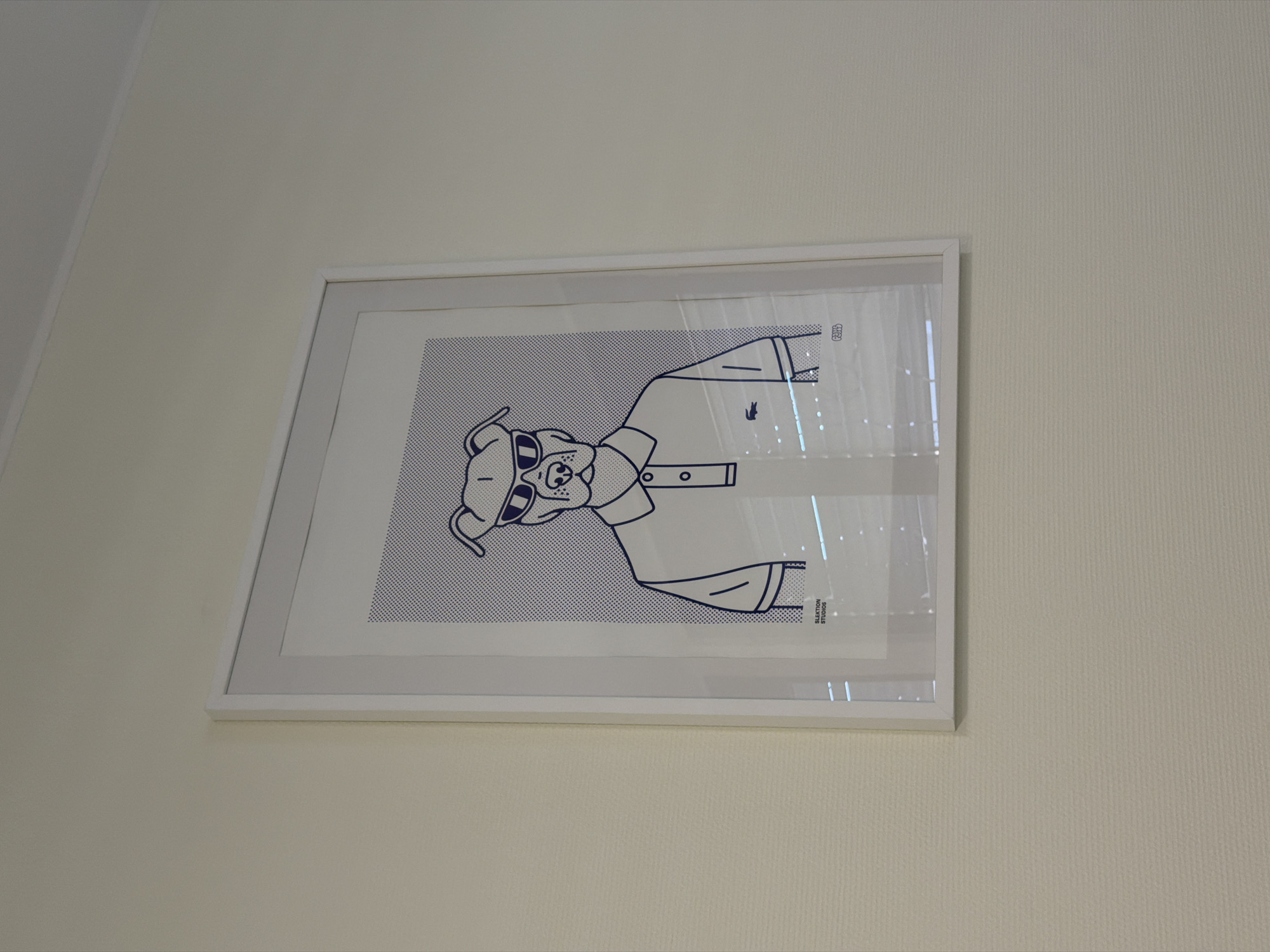 The image shows a framed cartoon picture of a dog wearing sunglasses and a shirt. The picture is hanging on a light beige wall. The frame is white. The style of the drawing is minimalist, with thick blue lines on a white background filled with small blue dots. The shirt has a small crocodile logo. The bottom right of the picture says "SLAKTION STUDIOS". The location appears to be a wall in a building in Esch-sur-Alzette, Luxembourg.