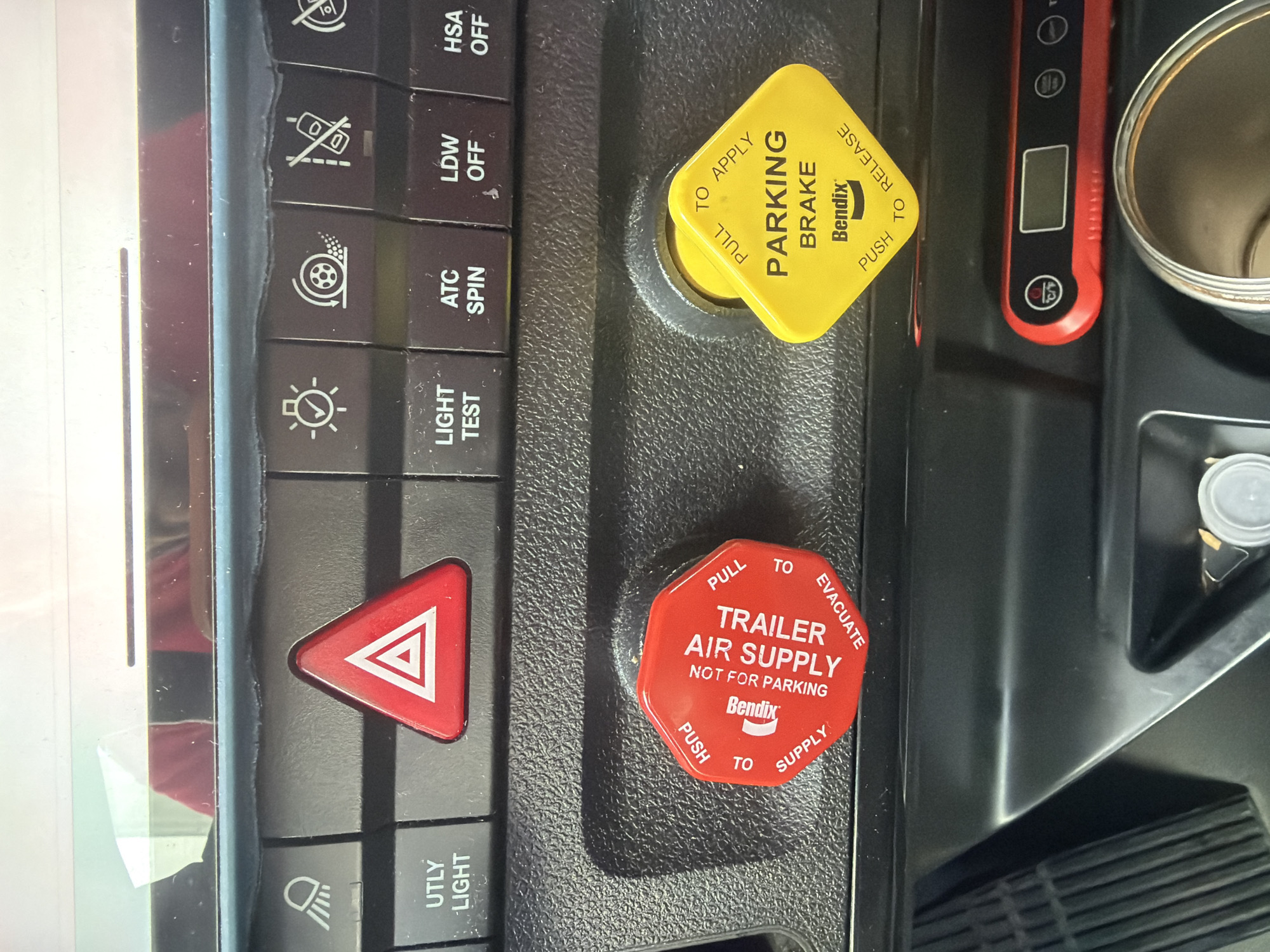 The image displays the interior of a truck's control panel, specifically focusing on the dashboard area. Prominently featured are two large, distinct control knobs: a red octagonal knob labeled "TRAILER AIR SUPPLY NOT FOR PARKING" with "PUSH TO SUPPLY" and "PULL TO EVACUATE" instructions, manufactured by Bendix. To its right is a yellow diamond-shaped knob labeled "PARKING BRAKE" with "PUSH TO RELEASE" and "PULL TO APPLY" instructions, also manufactured by Bendix. Above these knobs, a row of smaller buttons is visible, including a red hazard light button with a white triangle, and other buttons with symbols and text such as "UTLY LIGHT," "LIGHT TEST," "ATC SPIN," "LDW OFF," and "HSA OFF." The background shows a textured, dark surface typical of truck interiors, with a faint reflection of what appears to be a person in red clothing on the upper portion of the dashboard. The image is a close-up shot, capturing the detail of the controls. The setting is not identifiable as being in Lebanon, United States of America.