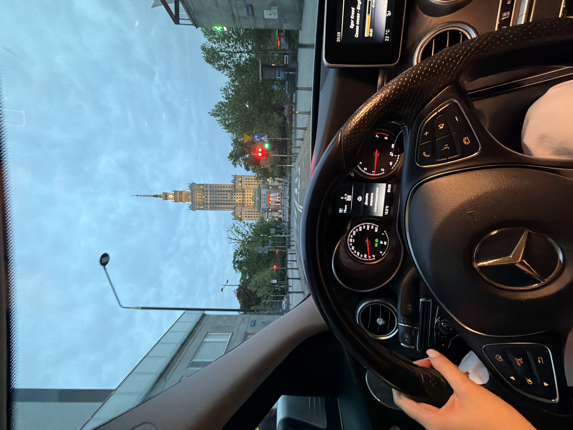 The image shows a view from inside a Mercedes-Benz car, presumably at a red light, as the instrument panel and the car's steering wheel are prominent in the foreground. A person's hand is visible on the steering wheel, but the rest of their body is not within the frame. Through the windshield, the Palace of Culture and Science in Warsaw, Poland, is clearly visible. Streetlights, trees, and buildings are also visible outside the car. The infotainment screen indicates that it is 20:28 (8:28 PM), and the music is playing "Самая самая" by Egor Kreed. The temperature inside the car is 22°C, while the outside temperature is 12.5°C.