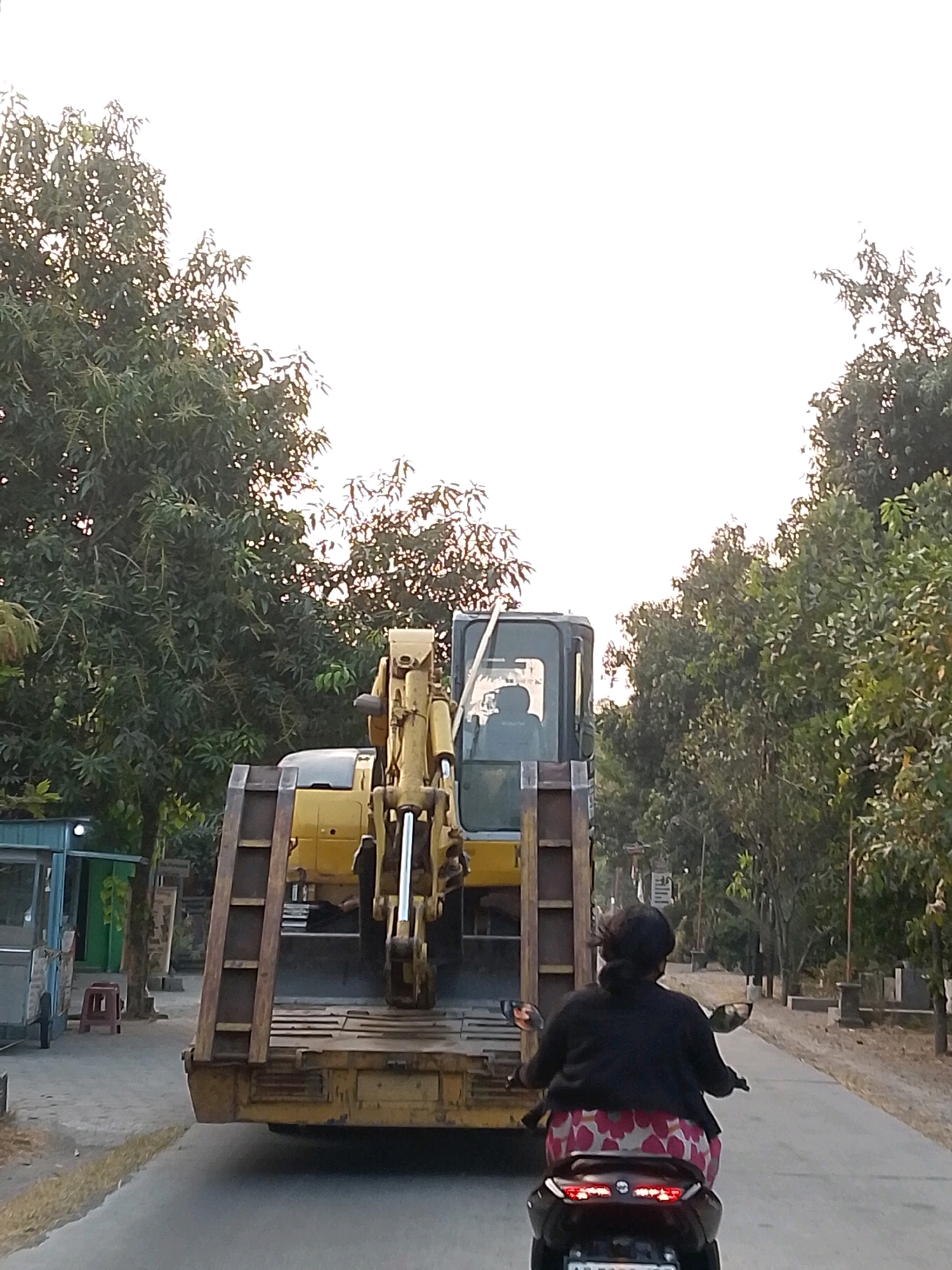 A low-angle, eye-level shot shows a yellow excavator on a flatbed trailer being driven down a road. The trailer is the primary focus, filling the center of the frame. A woman is riding a motorcycle in front of the trailer, obscuring part of it. The motorcycle is facing away from the camera. Trees line both sides of the road, and the sky is bright and overcast. To the left of the road, there is a blue and gray structure with a red stool in front of it. The scene appears to be in Semen, Indonesia.