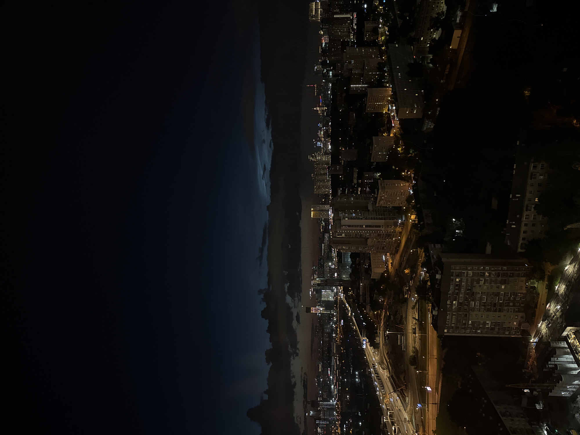 The image is a nighttime aerial view of a city on the right, with a vast, dark expanse on the left. The city is illuminated by countless lights from buildings and streetlights, creating a vibrant urban landscape. Various buildings, including what appear to be residential high-rises and commercial structures, are visible. Roads with flowing traffic lights weave through the cityscape.
The left side of the image is dominated by darkness, with a distinct band of clouds or storm front running horizontally, separating the city from the deep night sky. The clouds are illuminated from below by the city lights, creating a gradient effect from dark blue to lighter grays. The overall mood is dramatic, possibly capturing the transition into a storm or the edge of a weather system. The time of day is clearly night. There are no visible people or animals. The scene is set in an urban environment, likely a coastal city given the expanse of dark water or sky on the left. No text is visible in the image.