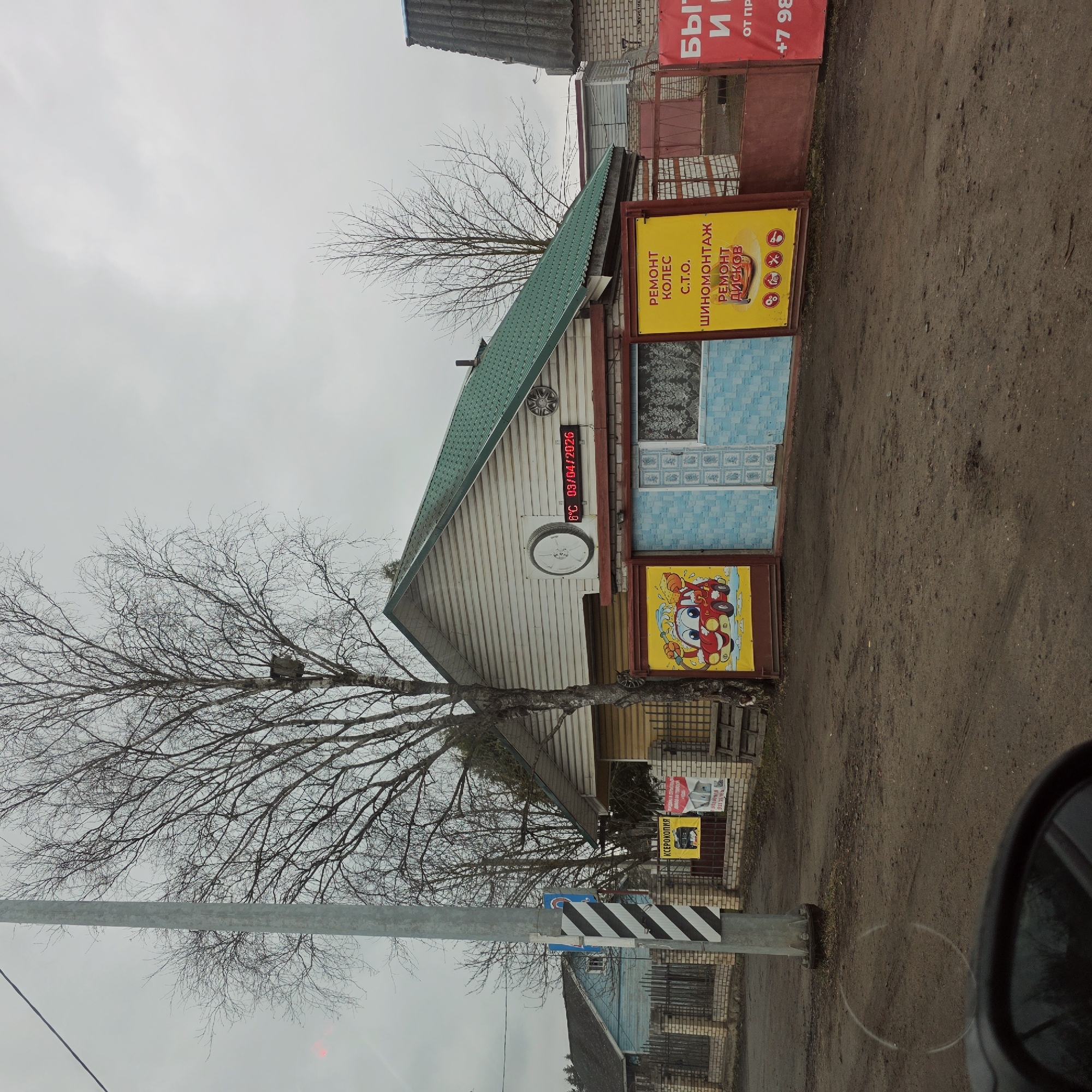 The photograph depicts an outdoor scene in what appears to be a rural or semi-rural area, likely in Volosovo, Russia, as indicated by the OCR text on the signage. The most prominent structure is a small, one-story building with a green roof and white wooden siding. It seems to be a repair shop, judging by the signs attached to the building advertising "Ремонт Колес" (tire repair), "Шиномонтаж" (tire fitting), "Ремонт Дисков" (wheel repair), and "C.T.O." (vehicle maintenance station). The temperature and date, "6°C 03/04/2026," are digitally displayed. Another sign featuring a cartoon car is present. A bare tree stands near the building, obscuring some of the view. A striped utility pole is in the foreground. In the background, other buildings are visible, including one that is partially brick. The ground is unpaved and appears to be dirt or gravel. The weather seems overcast. Part of a vehicle's side mirror is visible in the lower right corner of the frame. There are also signs with Russian text visible on the right.
