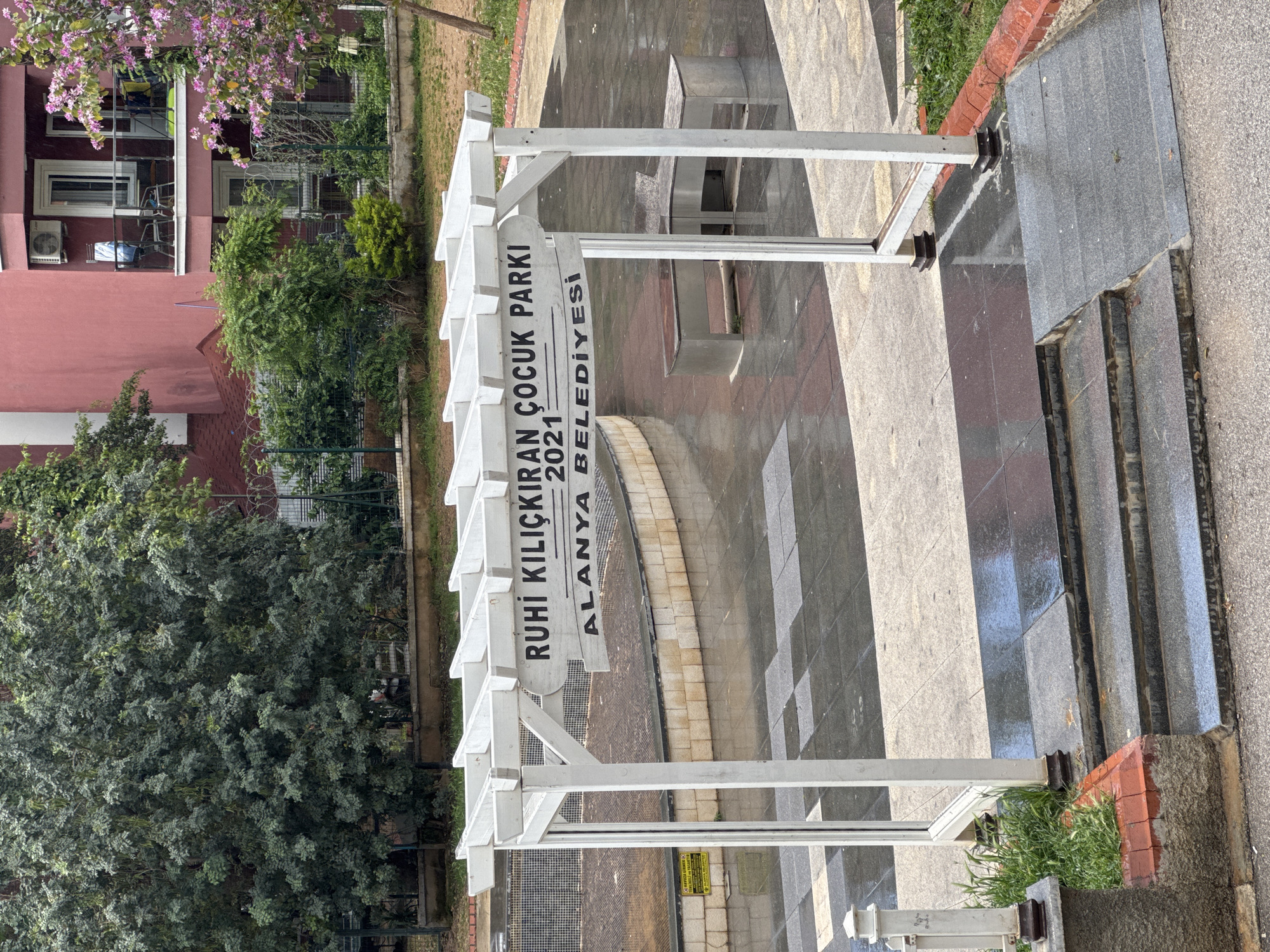 The image shows an entrance to a park. A white pergola stands at the entrance with a sign that reads "RUHI KILICKIRAN COCUK PARKI 2021 ALANYA BELEDIYESI". The park entrance features a tiled walkway with a checkered pattern, and stone steps lead into the park. A metal bench is visible in the background. Vegetation, including trees and grass, surrounds the park entrance. A building with red paint is partially visible in the background. The location is Alanya, Turkey.