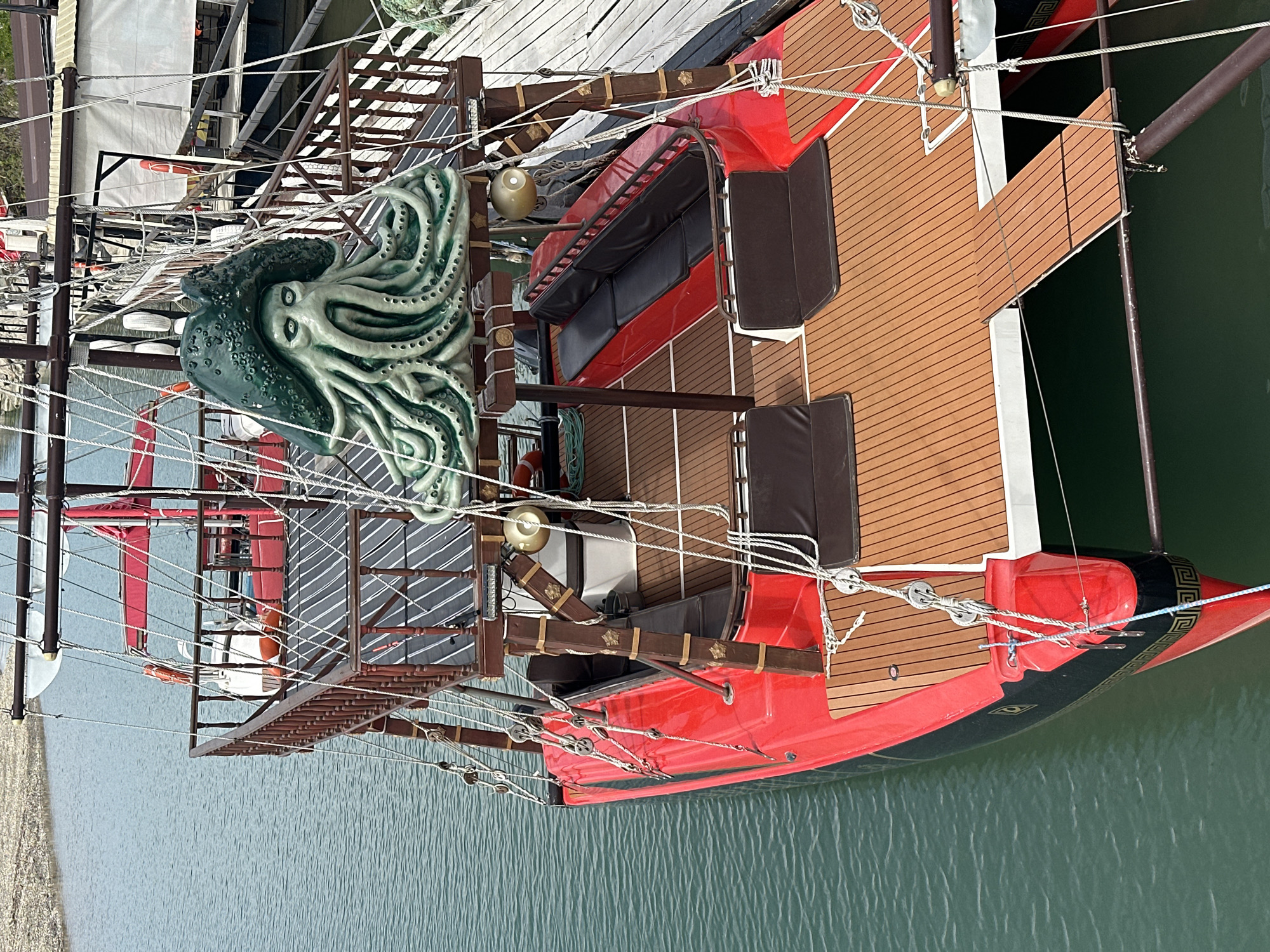 The image shows a pirate ship docked in the water, likely in the city of Novomikhaylovsky, Russia. The ship has a red and black hull, with a wooden deck and seating areas. A large green sculpture of an octopus adorns the front of the ship. Rigging and ropes are visible throughout the vessel. The water is calm and green. The ship is docked adjacent to a pier.