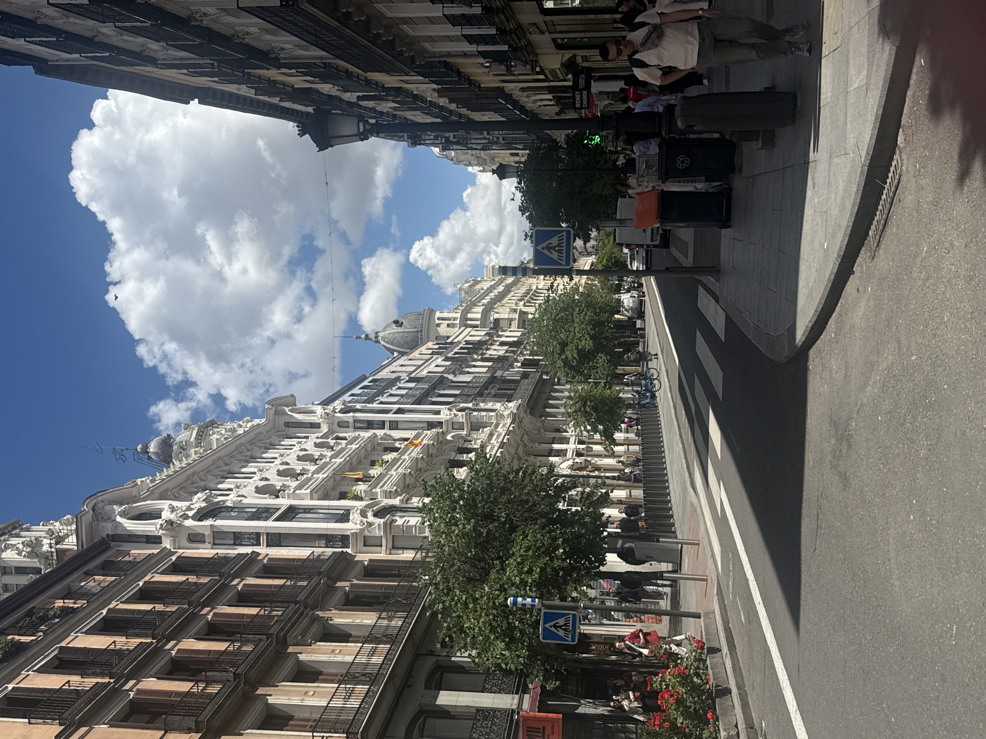 The image depicts a street in Madrid, Spain. The buildings on either side of the street exhibit classic European architectural styles. The sky is blue with fluffy white clouds. Pedestrians walk along the sidewalks on either side of the street. Some trees are visible along the sidewalk. There is a crosswalk marked on the street. There are street signs visible. The street appears to be clean and well-maintained. The image is taken at street level.