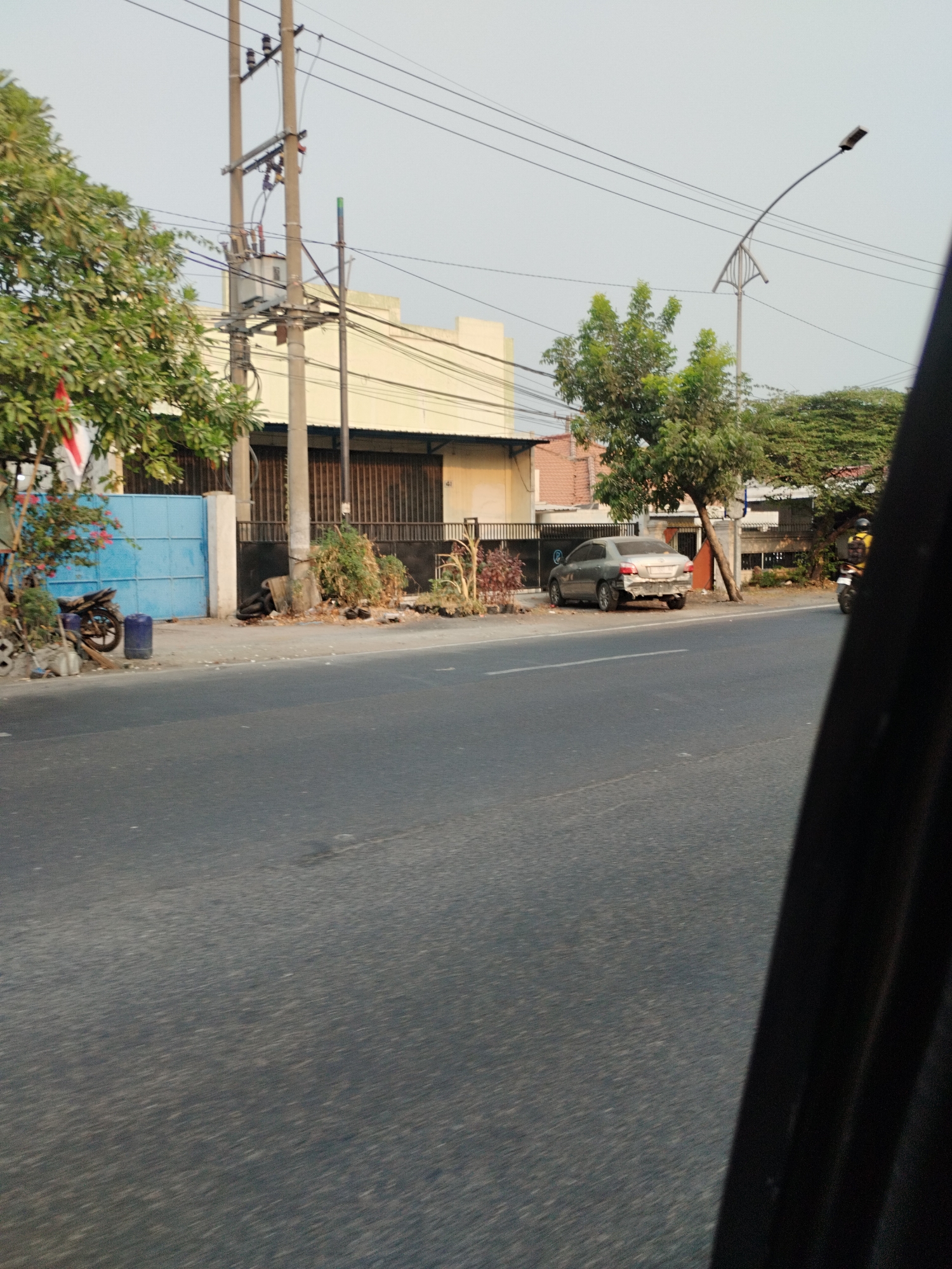 The image captures a street scene in Surabaya, Indonesia, likely during daylight hours given the illumination and clear sky. The perspective is from a moving vehicle, as indicated by the slight blur of the foreground asphalt and the dark, curved object on the right edge of the frame, which appears to be part of the vehicle's interior.
On the left side of the street, a light-colored building with a light yellow facade and a dark, corrugated metal roof is visible. A bright blue gate stands in front of this building, with a parked motorcycle to its left. A tall utility pole with multiple wires and a transformer is situated near the gate. Vegetation, including trees with green leaves and some flowering plants, borders the building and the sidewalk.
The street itself is paved with asphalt and has lane markings. On the right side of the street, there is a silver sedan with visible damage to its rear bumper. Further down the road, another utility pole and a street lamp with a curved arm are present, along with more trees. Another motorcycle, ridden by a person wearing a yellow shirt, is moving away from the camera. The overall environment suggests a typical urban street in Surabaya, with a mix of residential or commercial buildings, infrastructure, and ongoing traffic.
