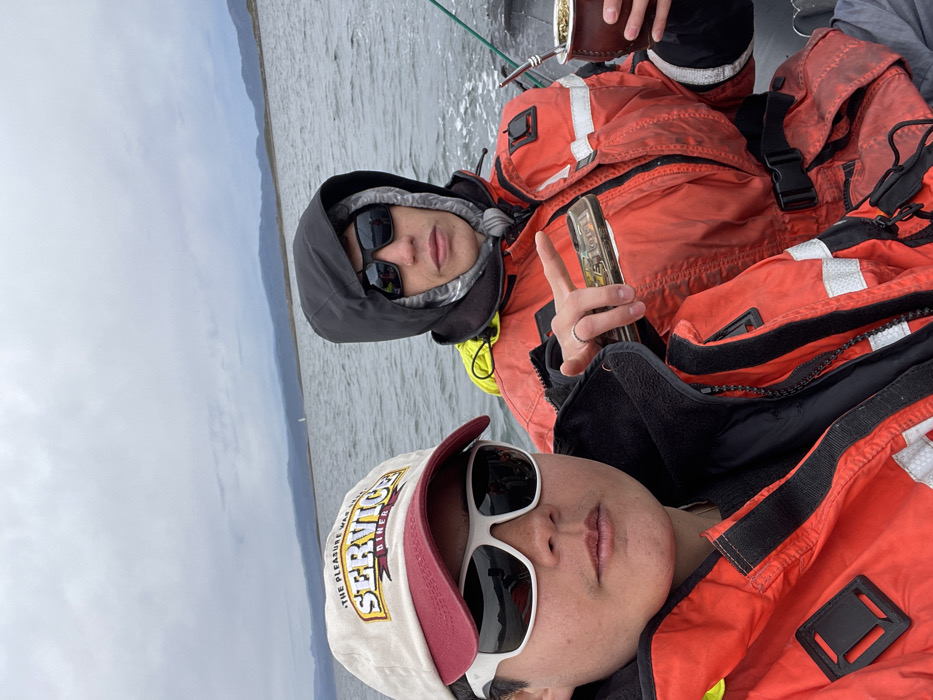Two individuals are featured in a selfie, taken outdoors on a boat on a body of water in Ushuaia, Argentina. Both are dressed in warm, protective gear, including orange and black life vests or heavy-duty outdoor jackets with reflective elements, indicating participation in a water-based activity in cool conditions.
The person in the foreground wears a light-colored baseball cap with a burgundy brim and white-framed sunglasses. Visible text on the cap includes "THE PLEASURE WAS OURS." and "SERVICE DINER". Their expression is neutral, partially obscured by the sunglasses.
Behind them, the second individual wears a dark hooded jacket with the hood up, paired with dark sunglasses. They are making a peace sign with their left hand, which also holds a rectangular, metallic-looking object, possibly a smartphone. A brown object, likely a mate gourd, is partially visible near their head.
The background shows a vast body of water with visible ripples, suggesting movement or wind. Distant mountains or hills stretch across the horizon under an overcast, cloudy sky, characteristic of the region. A small, faint white object, possibly another boat, can be seen on the water in the distance. The scene captures a moment of leisure or travel on the water, with the individuals prepared for a cool, possibly windy, environment.
