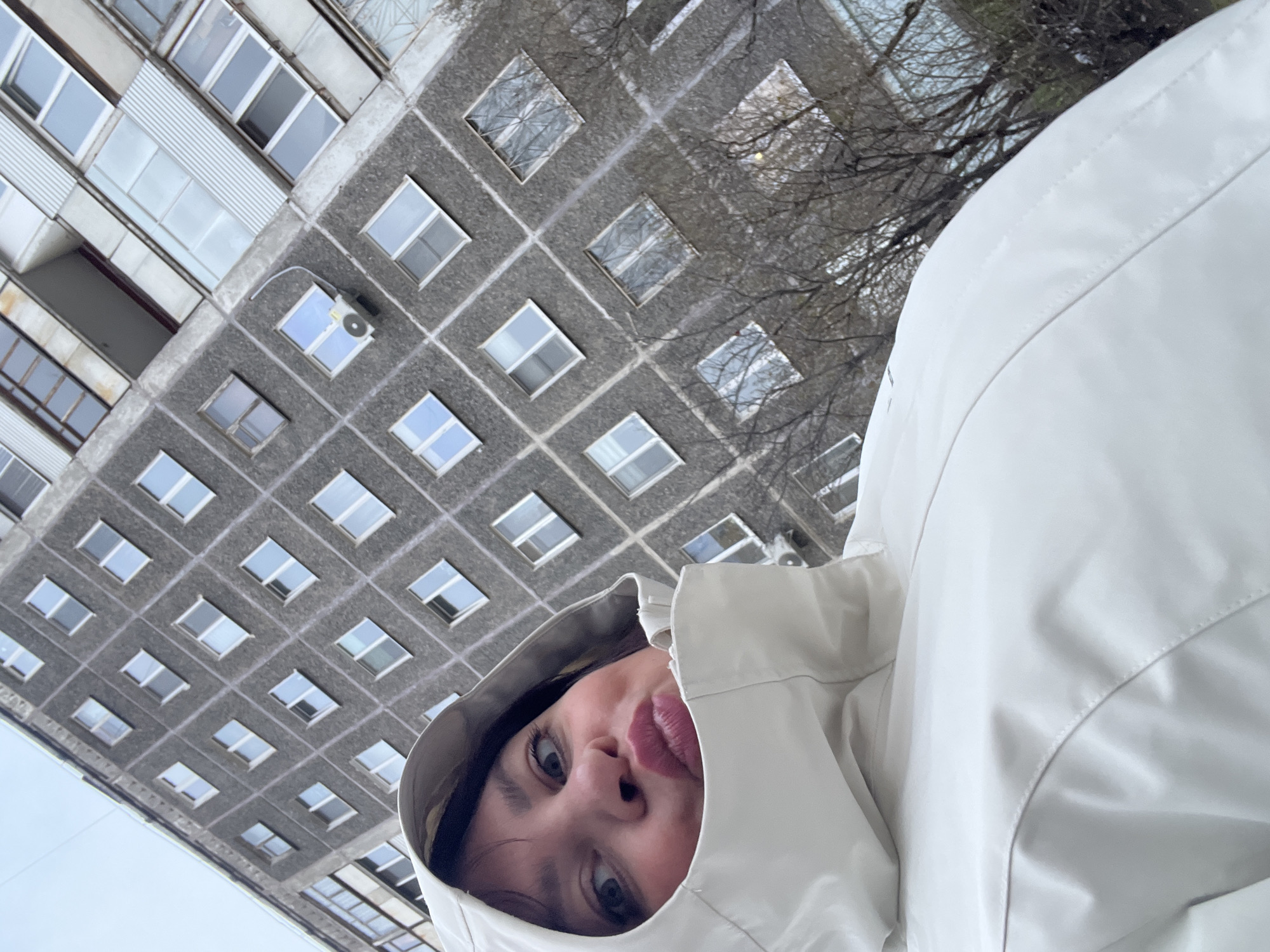 A tilted, low-angle shot captures a person in a white hooded jacket, appearing to look down at the camera, with a gray, multi-story building in the background. The building is characterized by numerous rectangular windows, suggesting an apartment complex or office building. Bare trees are visible to the right of the building. Based on the architecture of the building, this image is likely taken in Perm, Russia. The person's face is partially obscured by the hood, but eyes and lips are visible. The overall impression is an informal, perhaps spontaneous, snapshot.