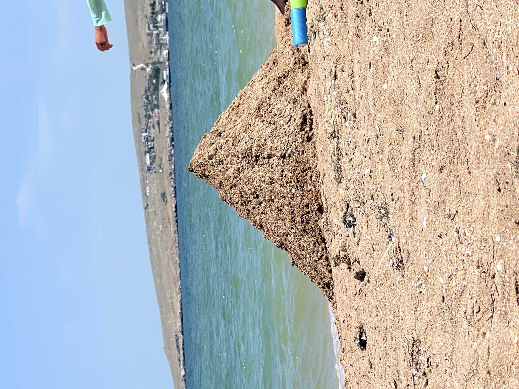 The image depicts a close-up view of a sandy beach with the sea in the background. A person's arm, clad in a light blue long-sleeved shirt, is visible extending from the upper left corner. They are reaching out, with their hand open. In the mid-ground, a triangular mound of sand, flecked with small shells and debris, dominates the scene. The sea is a vibrant turquoise, with gentle ripples indicating calm conditions. A distant shoreline features a town with buildings nestled along a hillside. The sky is clear and bright blue, suggesting a sunny day. The perspective is low and tilted, focusing on the texture of the sand and the contrast between the sand, sea, and sky. There is a small blue and green object visible in the upper right portion of the sand dune. The overall impression is of a bright, sunny day at a coastal location. The time of day appears to be daytime, given the bright sunlight and clear blue sky. The location cues, such as the town on the hillside and the sea, suggest a beach environment, possibly near a coastal settlement.