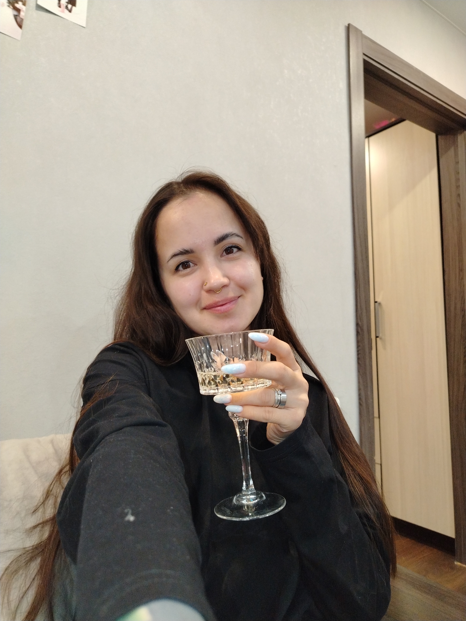 A medium shot depicts a woman indoors, holding a wine glass. She has long, dark brown hair and wears a black sweatshirt. Her left hand holds a glass of white wine, displaying light blue manicured nails. She has a nose ring and wears two rings on her ring finger. The background consists of a light gray wall, with a doorway revealing a closet or room with light-colored wood paneling. The location is Noviy, Russia.