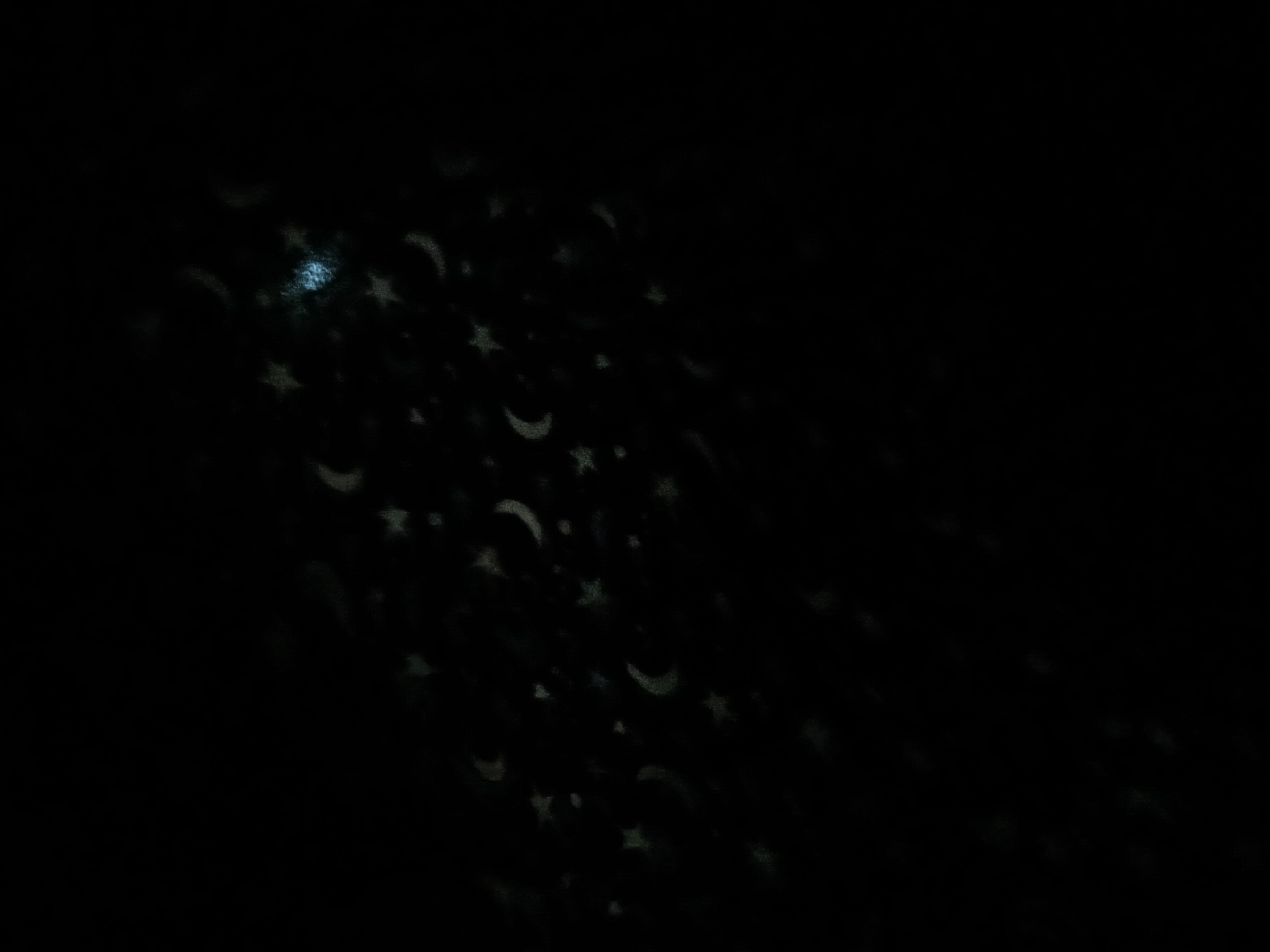 The image depicts a dark environment illuminated by projected patterns of stars and crescent moons. A faint light source is visible in the upper left corner. The scene is too dark to definitively determine the location as Altagracia, Venezuela. There are no people, subjects, objects, or actions clearly visible, besides the projected light patterns on a dark surface. The environment appears to be indoors.