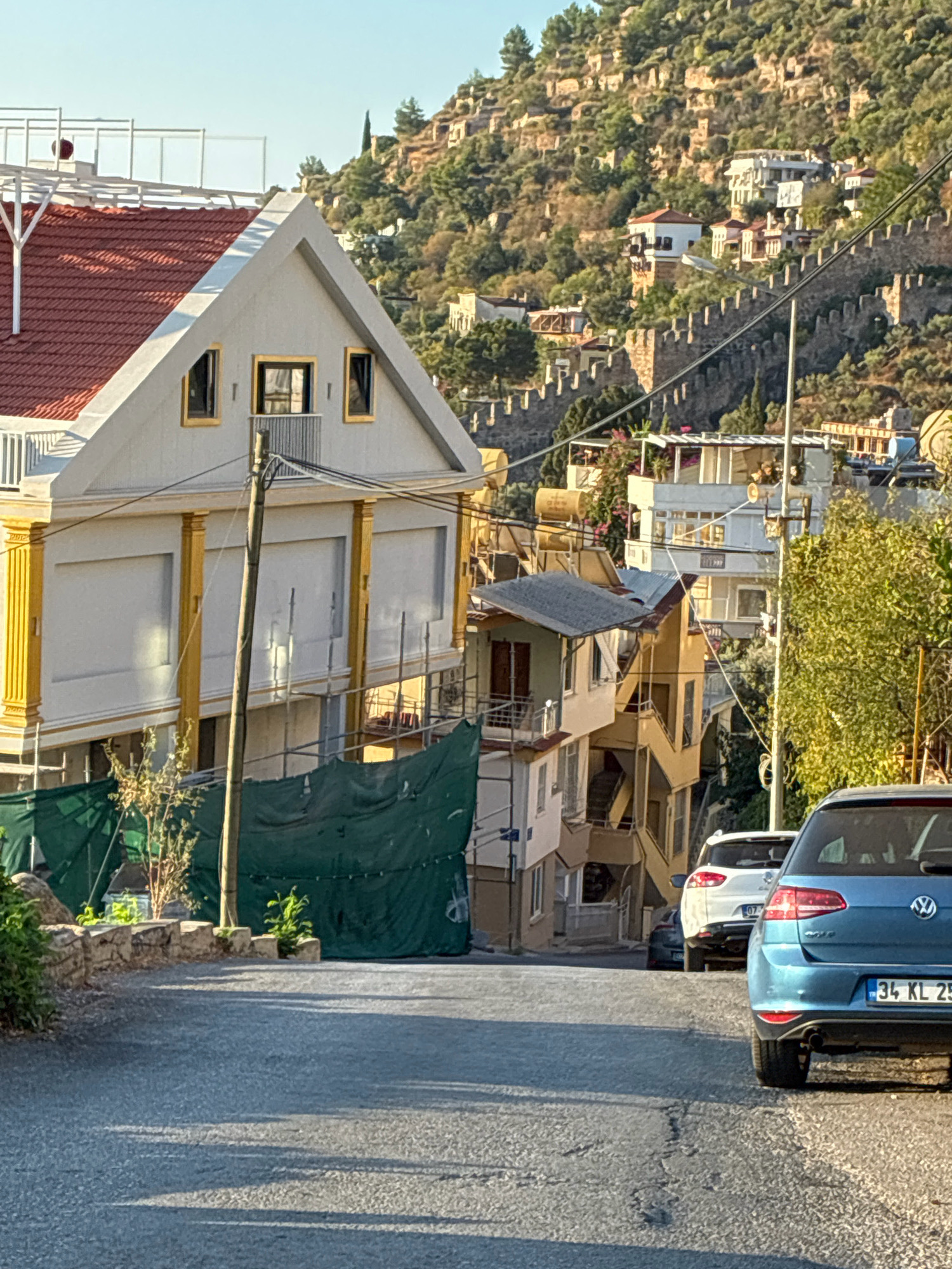 The image captures a steep, paved street in Alanya, Turkey, with a view of residential buildings and a historical fortress wall on a hillside. In the foreground, a blue Volkswagen Golf is parked on the right side of the street, facing away from the camera. To its left, a white SUV is also parked. The street slopes downwards, and shadows from the surrounding buildings and trees are cast across the asphalt.
On the left side of the street, a large, modern building with a red-tiled roof is under construction, partially covered by green tarpaulin. This building features white walls with decorative yellow columns. Further up the street, several older, multi-story houses are visible, built into the hillside. These buildings exhibit a mix of architectural styles, with balconies and terraces.
Dominating the background is a section of Alanya's ancient fortress wall, constructed from stone blocks, winding its way up the steep, rocky incline of the hill. Beyond the wall, more houses and buildings are scattered across the hillside, interspersed with trees and vegetation. The sky is clear and blue, indicating a sunny day. The overall scene depicts a dynamic urban landscape where historical architecture and modern development coexist on a challenging, hilly terrain.