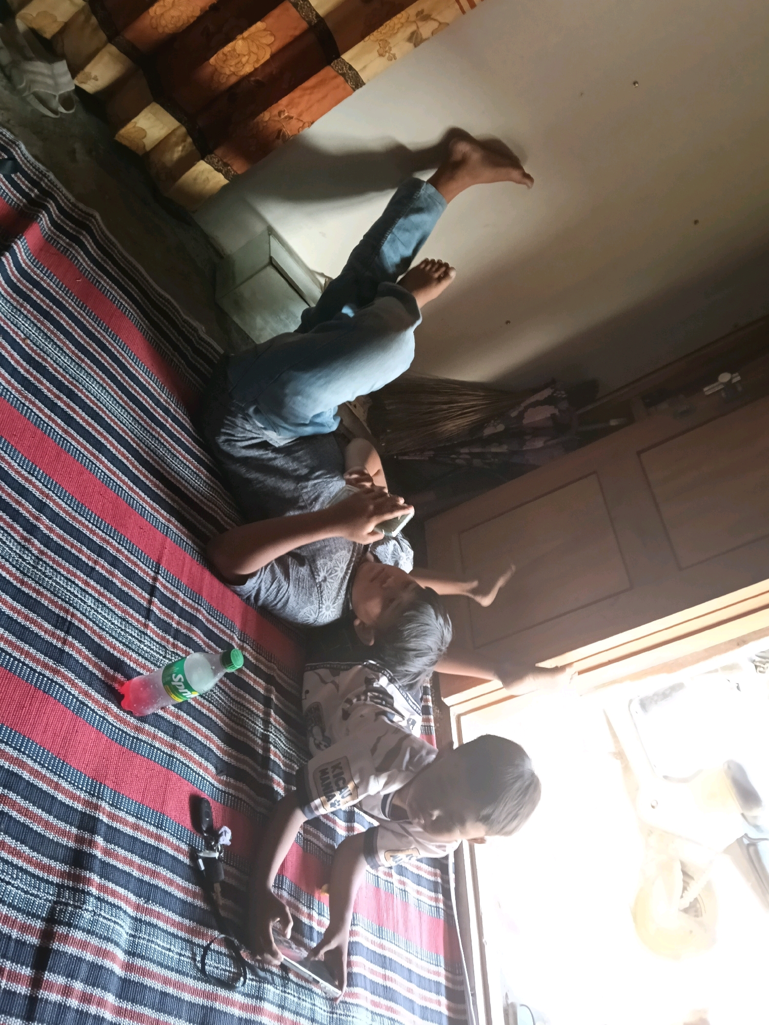 In Mendenrejo, Indonesia, two young boys are lying on a striped mat on the floor of a room. The boy on the left, wearing a grey t-shirt and blue jeans, is looking at his phone and holding it in his right hand. His legs are extended upwards. A bottle of Sprite with a pink liquid is next to him. The boy on the right, wearing a white t-shirt with a black and white pattern, is also looking at his phone, which he holds in his hands. His body is positioned in a way that suggests he is also lying down. The room appears to be simply furnished, with a wooden cabinet visible in the background and a window letting in bright light. The overall scene suggests a casual indoor activity.
