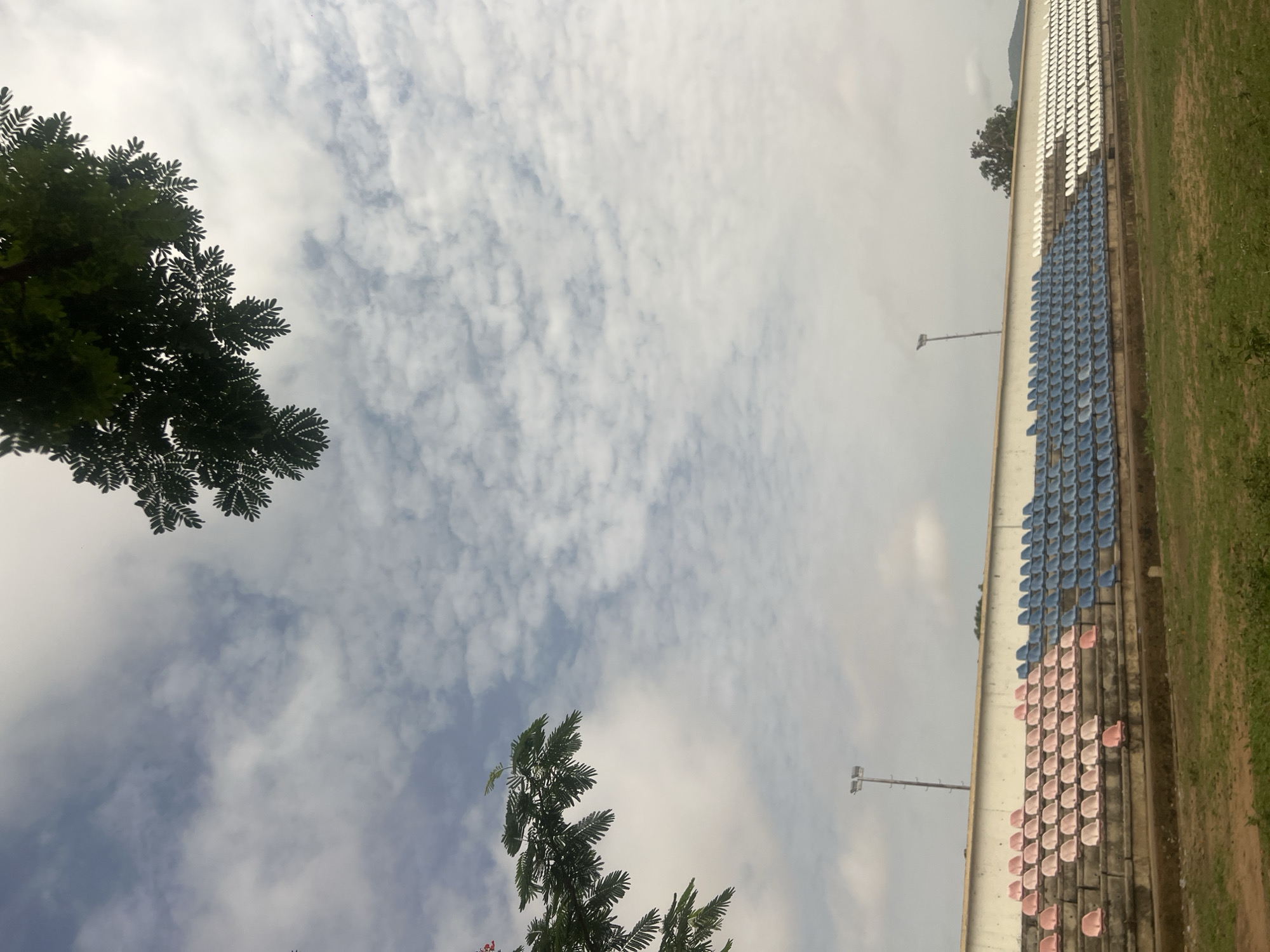 The image shows stadium seating in Jalingo, Nigeria, under a cloudy sky. The seating is tiered, with sections colored white, blue, and pink. Green grass borders the structure on one side. Sparse trees are visible in the periphery. Two stadium lights stand nearby.