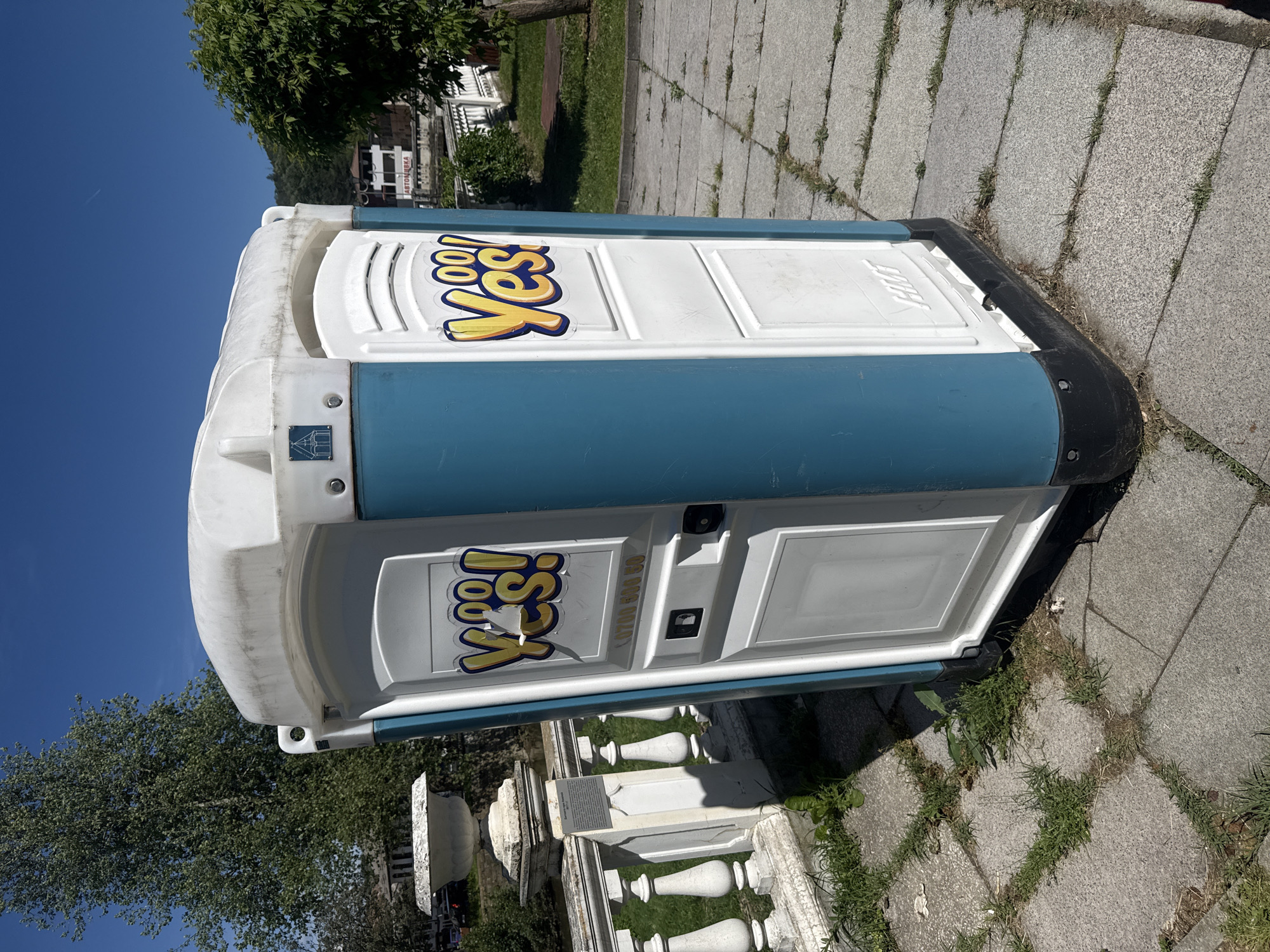 A portable toilet is located on a paved sidewalk in Tryavna, Bulgaria. The toilet is white with a blue stripe and has a "Yes!" logo on the side and door. The door of the toilet is slightly open. The sidewalk is made of gray stone slabs with grass growing in between the cracks. In the background, there is a white stone railing, trees, a building, and a blue sky.