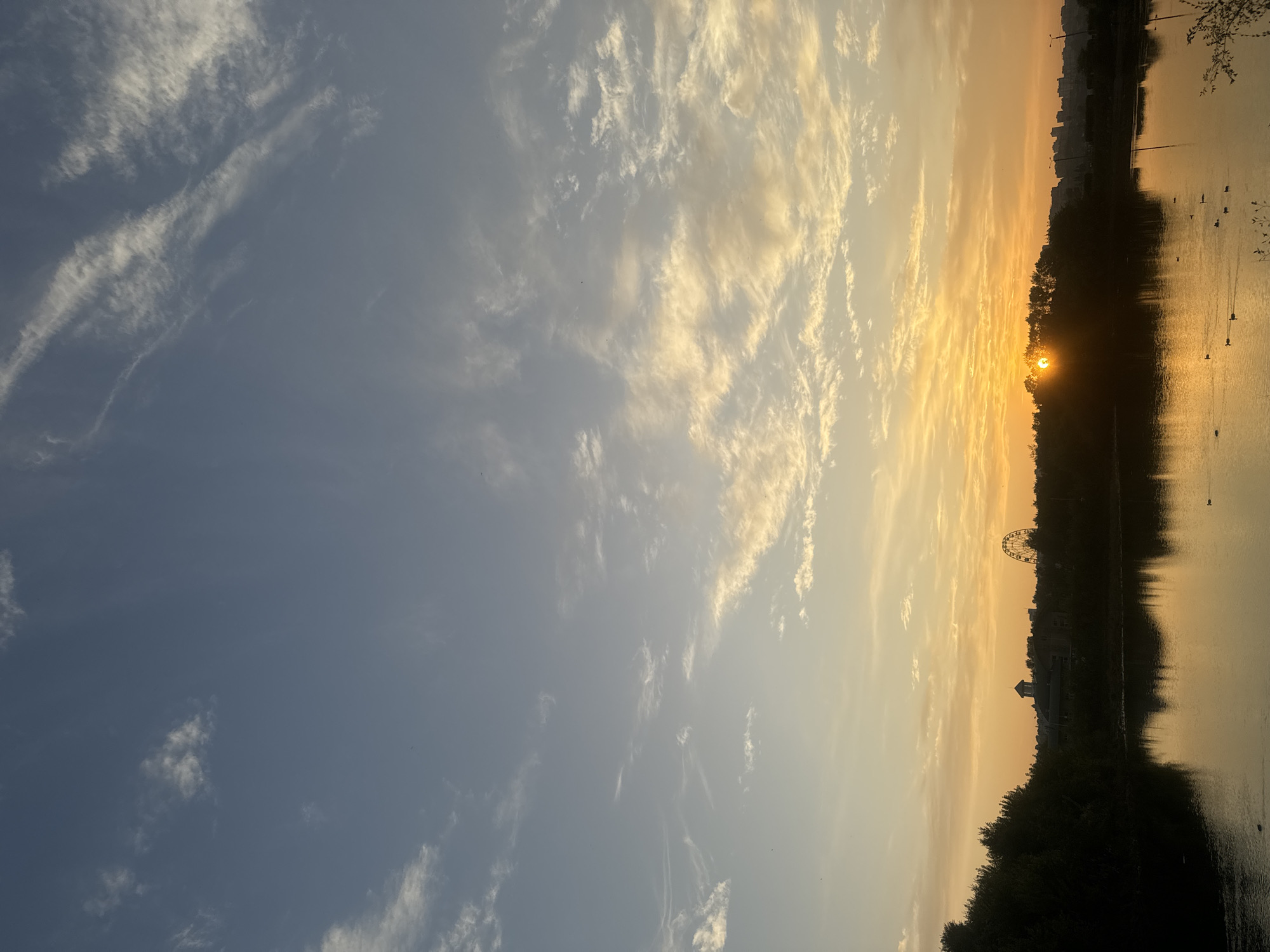 The image depicts a serene sunset over a body of water, likely a river or lake, in Irkutsk, Russia. Silhouetted trees line the left bank, with a Ferris wheel and buildings visible in the distance. The sky is a gradient of blue to warm orange, with wispy clouds catching the last rays of the sun. The water reflects the colors of the sky and the trees, creating a mirrored image. A few ducks are visible swimming on the water's surface. The overall atmosphere is peaceful and tranquil.
