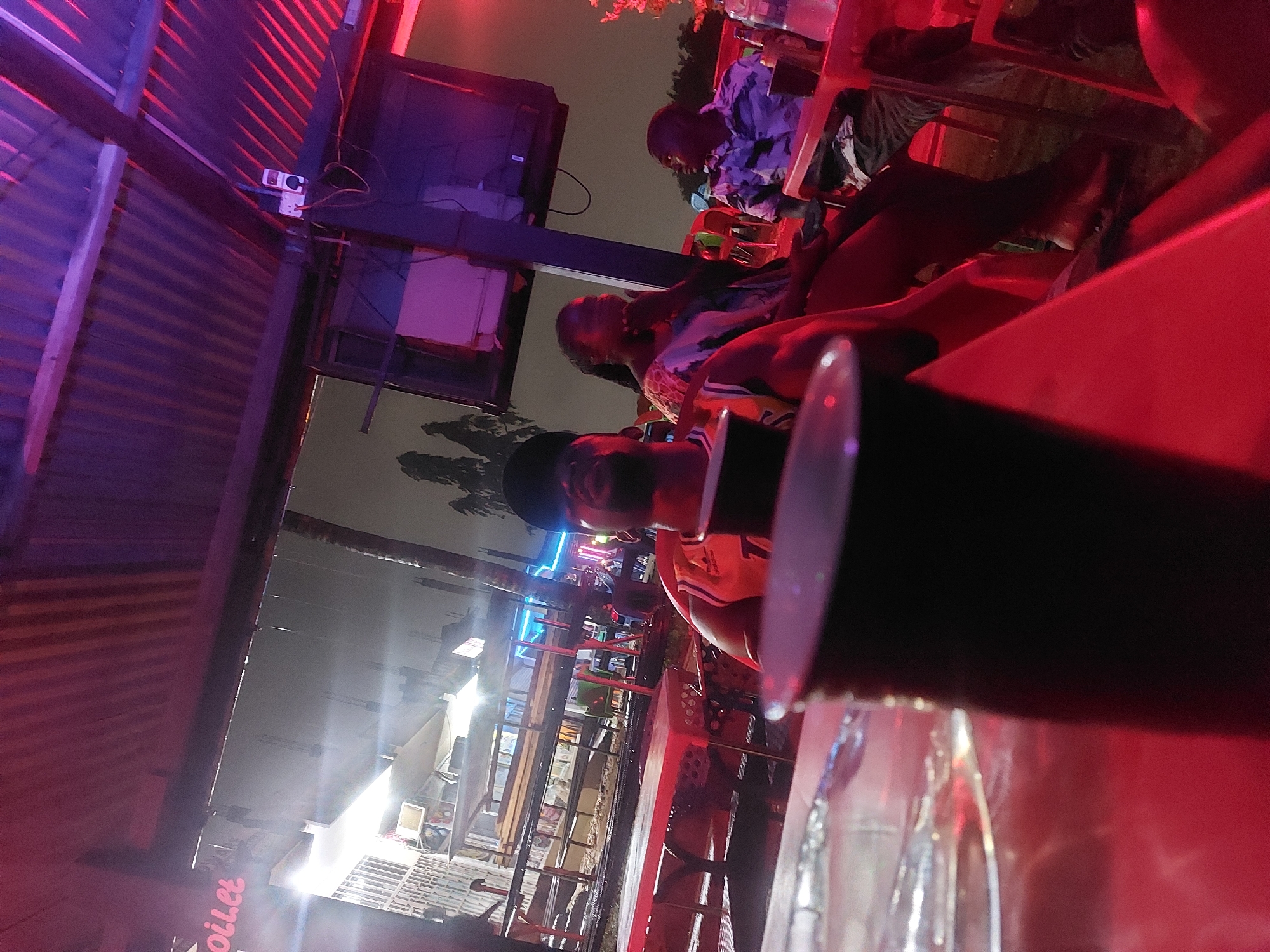 The image shows a night scene in Dar es-Salaam, Tanzania. Multiple people are seated at tables, possibly in an outdoor bar or restaurant. Red and blue lights illuminate the area. A surveillance camera is attached to a corrugated metal structure. A table with glasses and a cylindrical object is in the foreground.