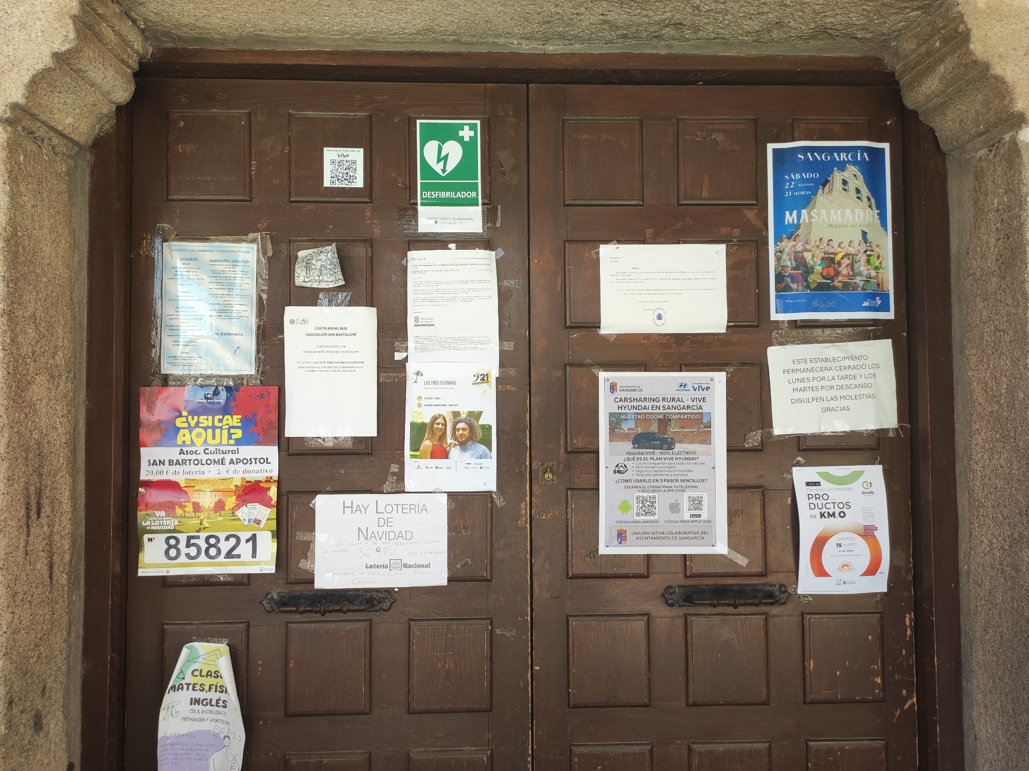The image depicts a weathered, wooden double door with numerous posters and flyers attached to its surface. The doors are set within a stone archway, suggesting an entrance to a building. The posters advertise various events, lotteries, and services, including a "Masa Madre" event in Sangarcía, a "Car Sharing Rural" service, a "Hay Lotería de Navidad" (Christmas Lottery), and classes in English, Mathematics, and Physics. One poster indicates that the establishment will be closed on Monday afternoons and Tuesday mornings for rest. A green defibrillator sign is also visible on the left door. The overall scene conveys a sense of community activity and local information dissemination within the town of Sangarcía, Spain.