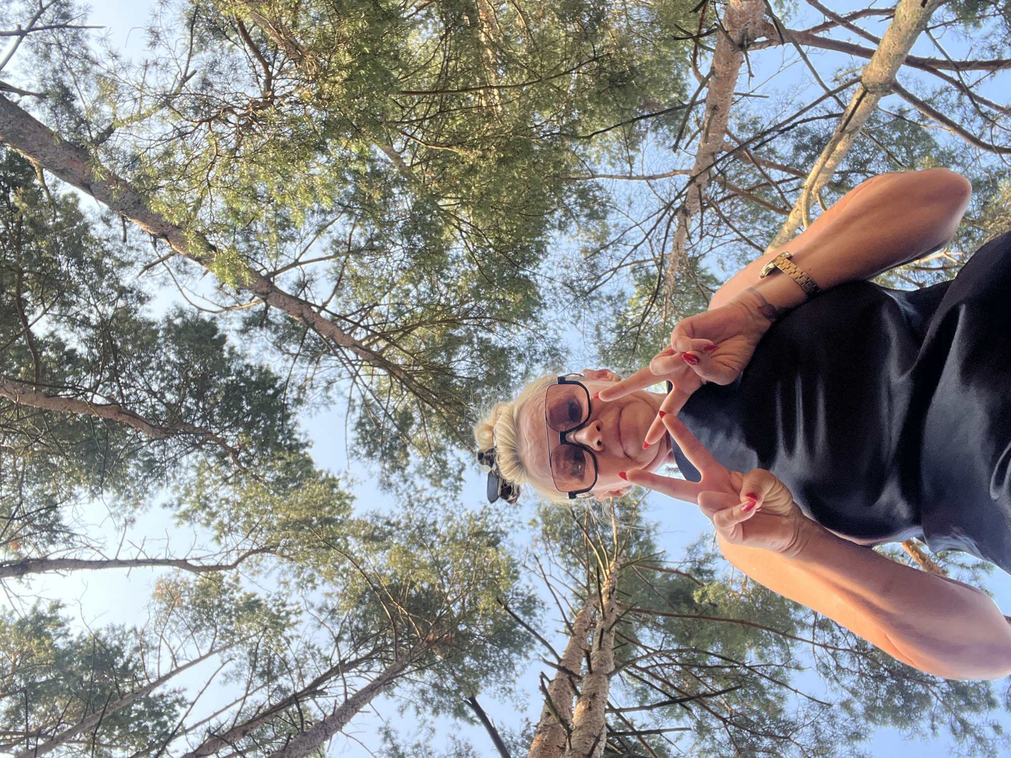 An extreme low-angle, upward-facing medium shot captures a blonde woman with pink-tinted sunglasses and bright red fingernail polish making a peace sign with her left hand and the index finger of her right hand extended upwards. She is wearing a black sleeveless top and a gold watch on her left wrist. Her blonde hair is pulled up into a bun with a leopard print accessory. The background consists of a clear blue sky with the branches and needles of several coniferous trees extending across the frame. The woman is positioned in the foreground, with her face tilted upwards towards the camera. The scene appears to be outdoors, possibly in a park or forest, given the presence of the trees. The location is identified as Kleosin, Poland.