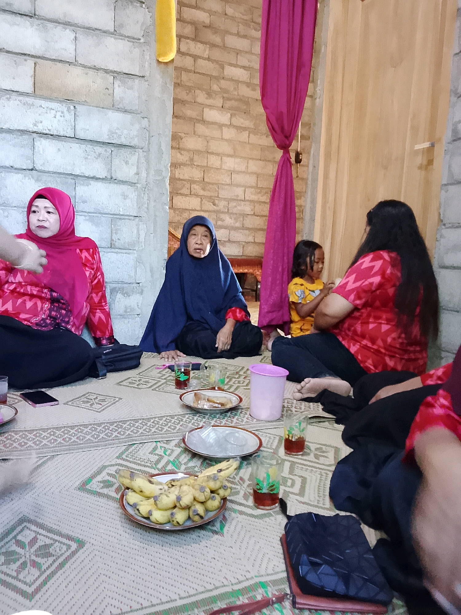 In Kradenan, Indonesia, a group of individuals is gathered on the floor of a room with brick walls. The room is furnished with a woven mat covering the floor, on which several objects are placed. In the foreground, a plate of bananas is centrally located. To the left of the bananas, a pink cell phone is visible. Further to the left, a woman wearing a magenta headscarf and a red, patterned long-sleeved shirt is seated. To her right, an elderly woman in a dark blue headscarf and a dark blue garment is also seated. In the middle ground, a young girl in a yellow t-shirt is seated next to a woman with long dark hair, who is wearing a red, patterned long-sleeved shirt. Two glasses filled with a reddish liquid and ice are placed on the mat, along with a pink plastic container and some plates. Two dark-colored bags and a brown wallet are also visible on the floor. The background features a partition made of light-colored wood and a draped magenta fabric. The overall atmosphere appears to be informal and relaxed.