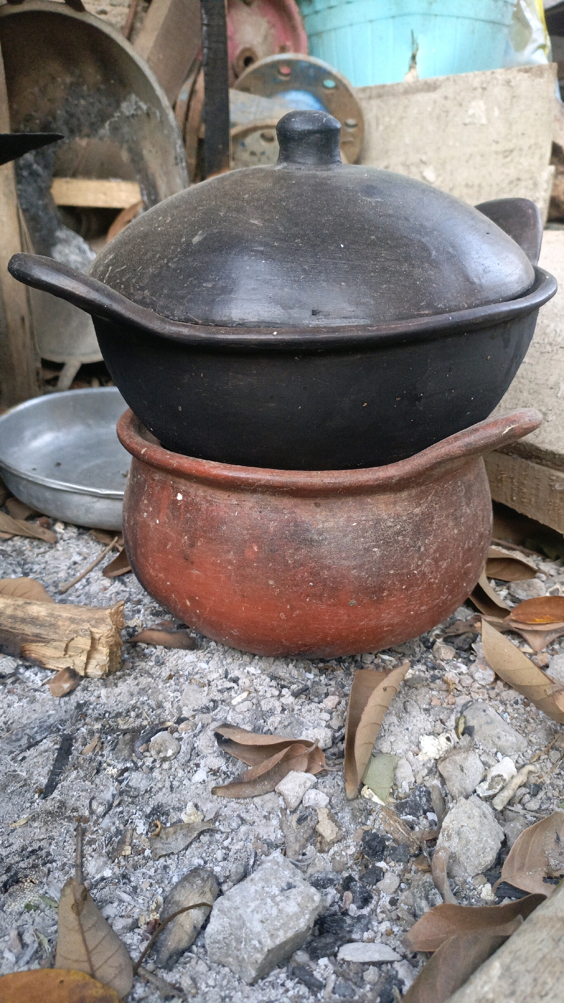 In Taliwang, Indonesia, a close-up, eye-level shot captures two clay pots stacked on a bed of ash and debris. The top pot is black and has a lid with a knob. It rests on a reddish-brown pot with handles that curve upwards. The surrounding ground is covered in ash, small stones, dried leaves, and pieces of wood. In the background, blurred objects suggest an outdoor setting, possibly near discarded items, with hints of metal and plastic containers. The scene appears to be a still life composition of cooking utensils in an outdoor, perhaps neglected, environment.