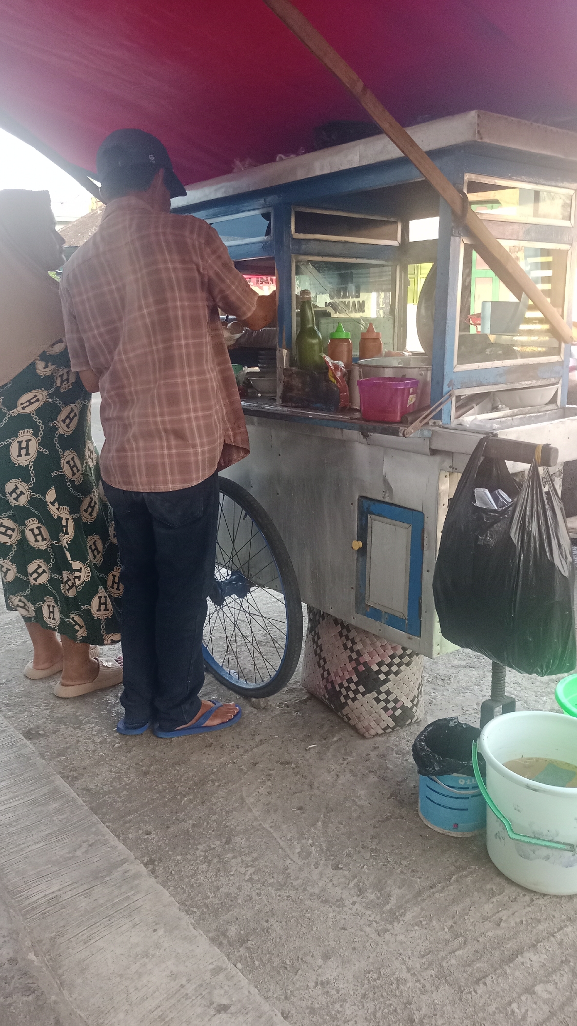The image depicts a street scene in Karyamandala, Indonesia, focusing on a food cart. Two individuals are partially visible. On the left, a person wearing a hijab and a patterned green skirt stands near the cart. To their right, a man in a plaid shirt and dark pants is interacting with the cart. The food cart itself is a mobile unit with a blue and silver facade, a red awning, and a large spoked wheel. It is stocked with various items, including bottles of condiments, containers, and what appears to be food preparation items within the glass display. A black plastic bag hangs from the cart, and a woven basket sits beneath it. The ground is paved with concrete. The overall lighting suggests it is daytime.
