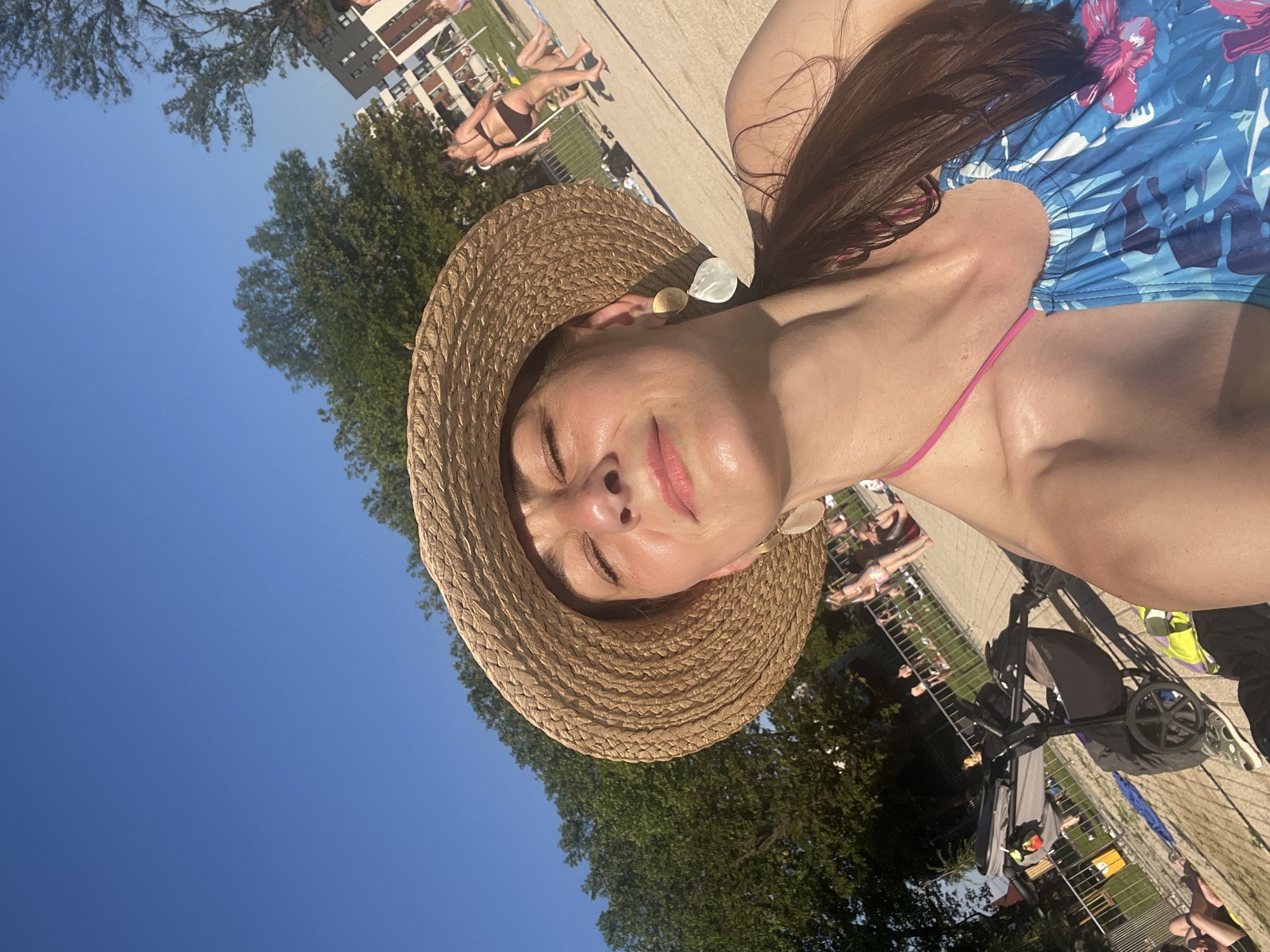 A woman is lying on her back with her eyes closed, wearing a straw hat and a floral swimsuit. She is in an outdoor setting, likely a poolside area, with a bright blue sky overhead. In the background, other people can be seen sunbathing and relaxing. There is a stroller visible, suggesting a family outing. The lighting indicates it is daytime, likely sunny given the shadows and the relaxed, sun-soaked appearance of the woman. The overall mood seems peaceful and leisurely. There is no visible text in the image.