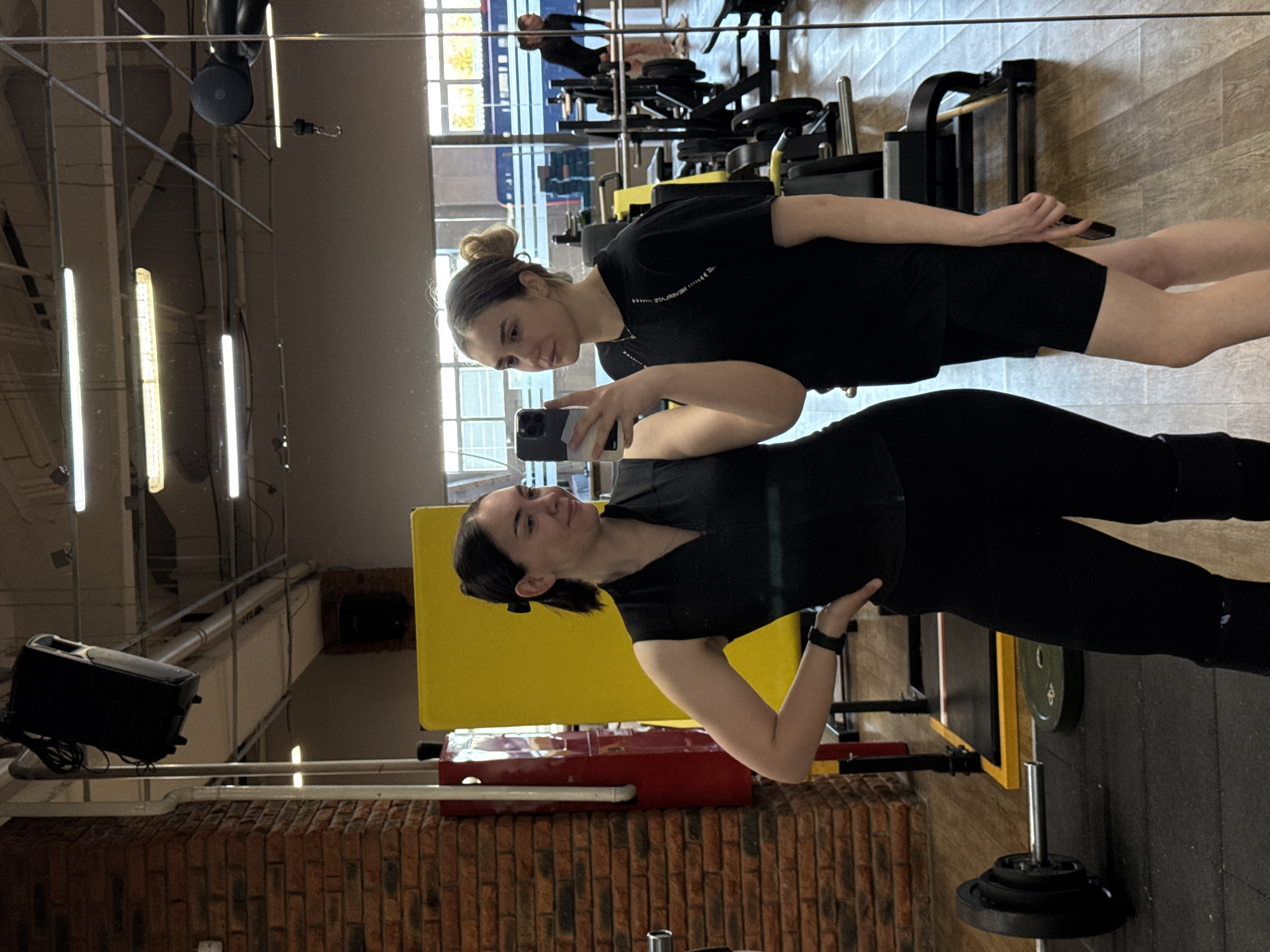 Two women are visible in a gym, reflected in a large mirror. One woman is holding a phone and appears to be taking a selfie, with the other woman in the frame. The woman taking the selfie is wearing black leggings and a black top, while the other woman is wearing black shorts and a black t-shirt. Gym equipment is visible in the background and foreground. The location is Chaikovsky, Russia.