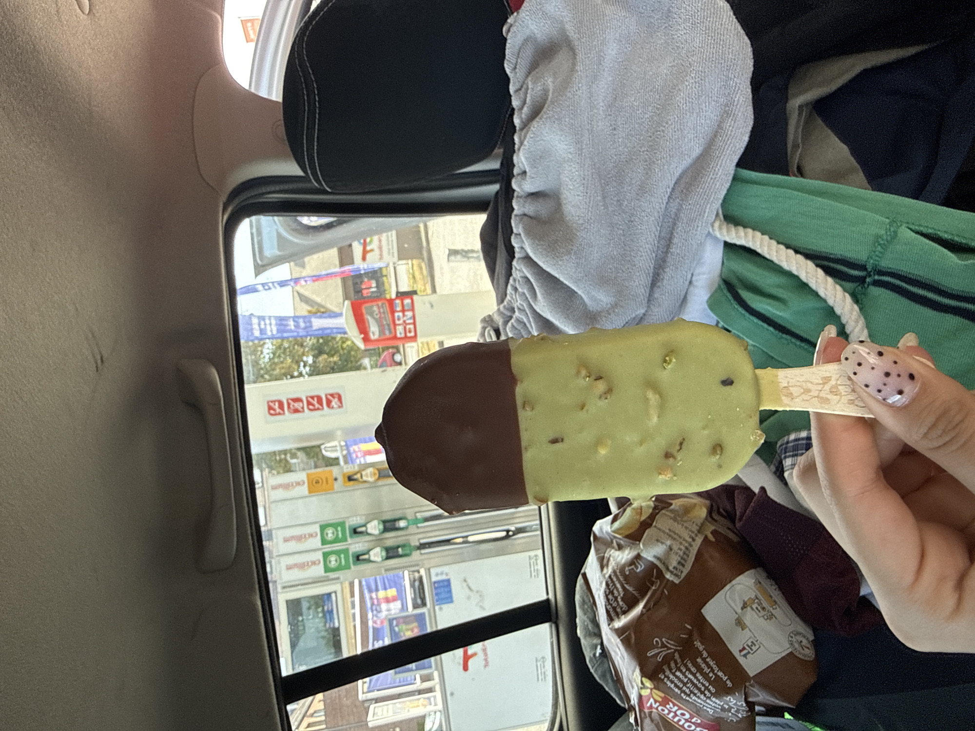 A person's hand with black polka dot nail art holds a chocolate-covered ice cream bar in the passenger seat of a vehicle. The ice cream bar has a dark chocolate top and a light green, pistachio-flavored bottom with visible pistachio pieces. In the background, through the car window, a Total gas station with multiple pumps and signage is visible. The gas station is located on Rue de la Carme in Mont-Saint-Guibert, Belgium. The car interior is cluttered with various items, including a bag of chips, a grey towel, and green and blue clothing. The sky is overcast.