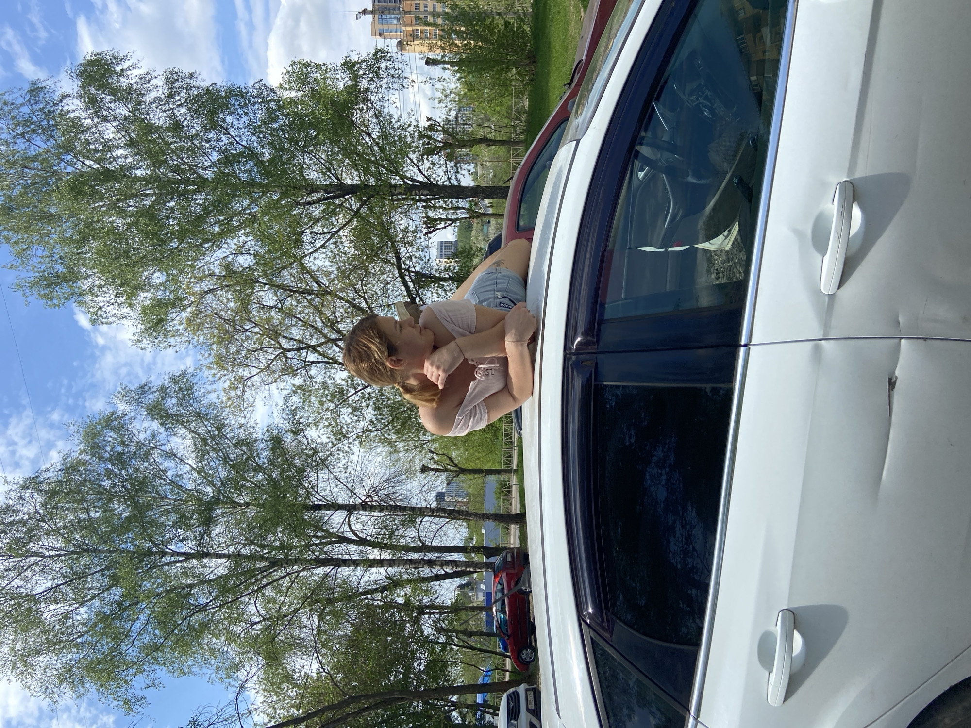 A young woman is leaning out of the open driver's side window of a white car in Ryazan, Russia. She has light brown hair and is wearing a light pink top and denim shorts. She is resting her head on her hand and looking out at the trees. The car is parked on a street lined with trees. In the background, there are apartment buildings. The sky is blue with some clouds.