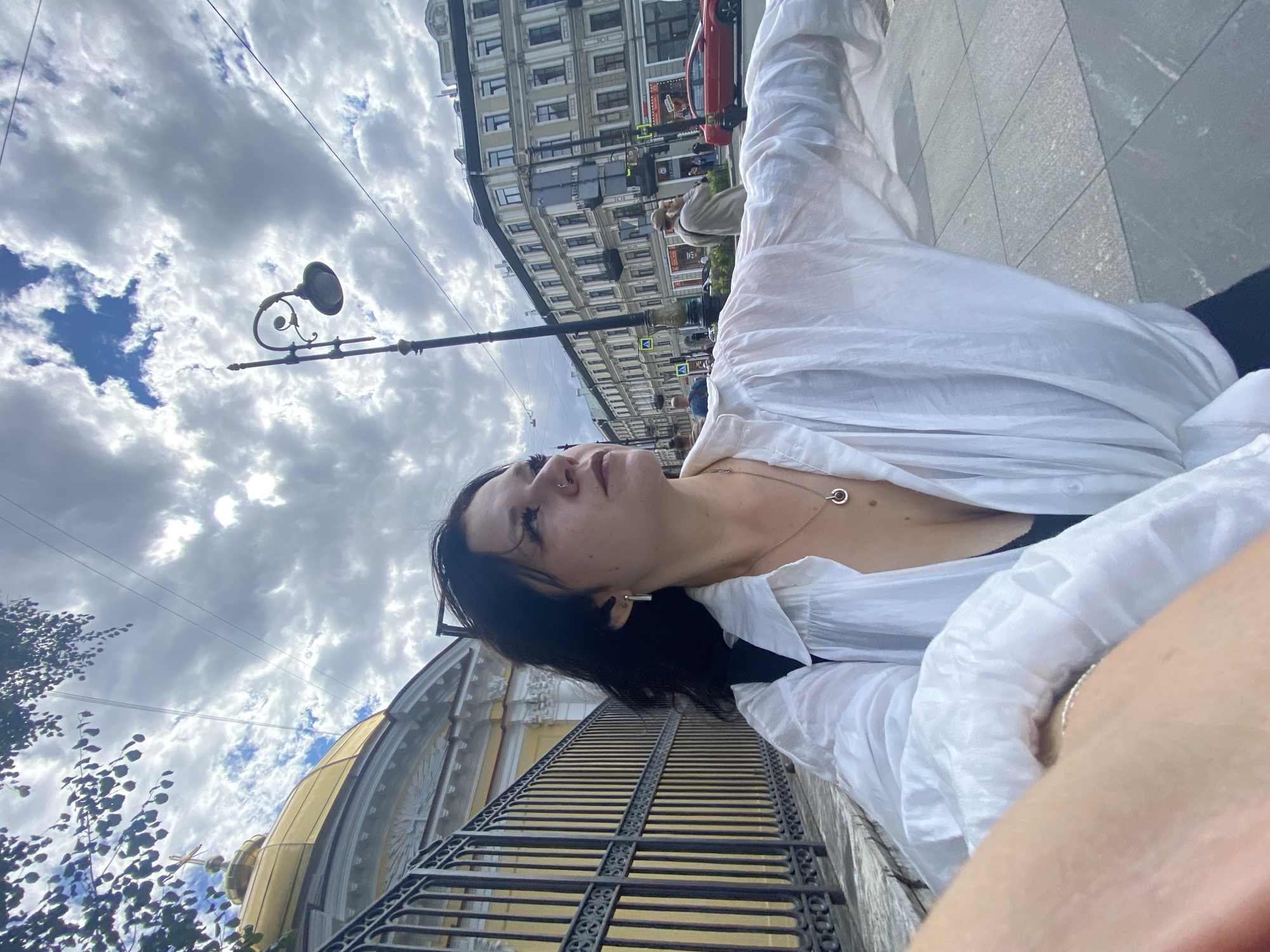A woman with dark hair, wearing a white open button-up shirt over a black top and a necklace, stands on a sidewalk in Saint Petersburg, Russia. She is looking to her right, with her left arm extended towards the camera. Behind her, a gold-domed building with a cross on top is visible to her left, separated by a black metal fence. A lamppost stands in the middle ground. In the background, multi-story buildings line a street with people, cars, and traffic signals. The sky is filled with dramatic, cloudy formations.