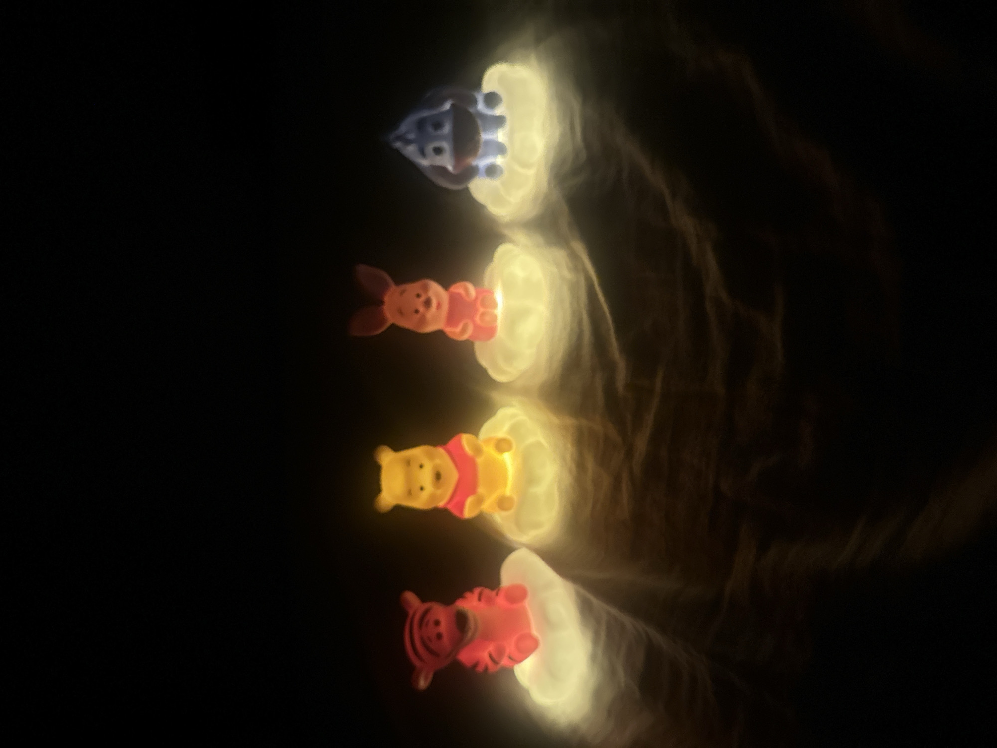 Four illuminated figurines are aligned vertically in a dark environment. From top to bottom, the figurines appear to be: Eeyore (blue), Piglet (pink), Winnie the Pooh (yellow), and Tigger (orange). Each figurine sits atop a glowing, white or yellowish light source. The background is primarily dark, with some subtle brown shadows visible. The image does not provide any information to associate the scene with the city of Votkinsk, Russia.
